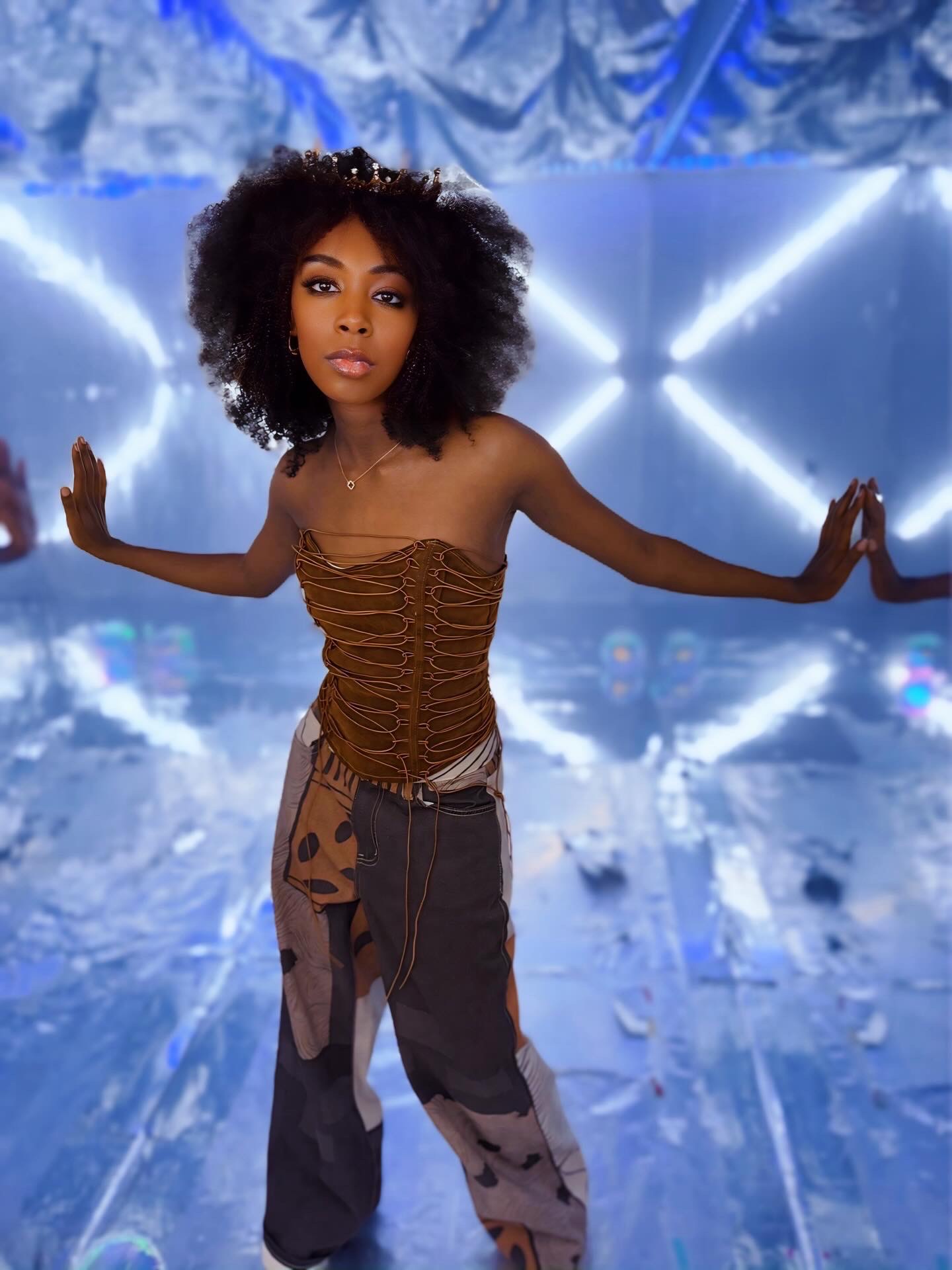 Young woman with curly hair stands with arms outstretched in a futuristic setting with blue lighting and geometric shapes.