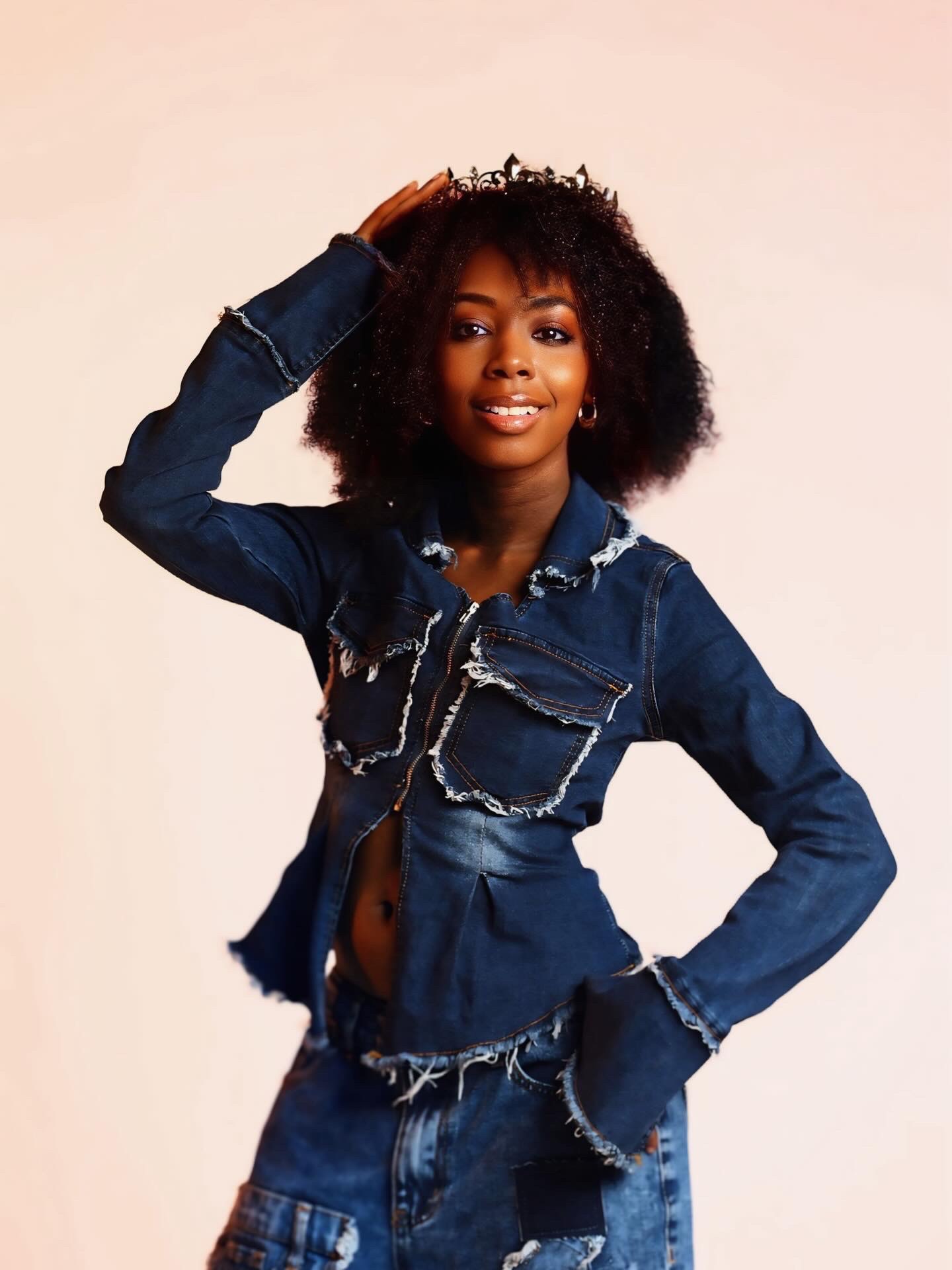 Young woman with curly hair wearing a denim jacket and jeans, smiling, posing with one hand on her head.