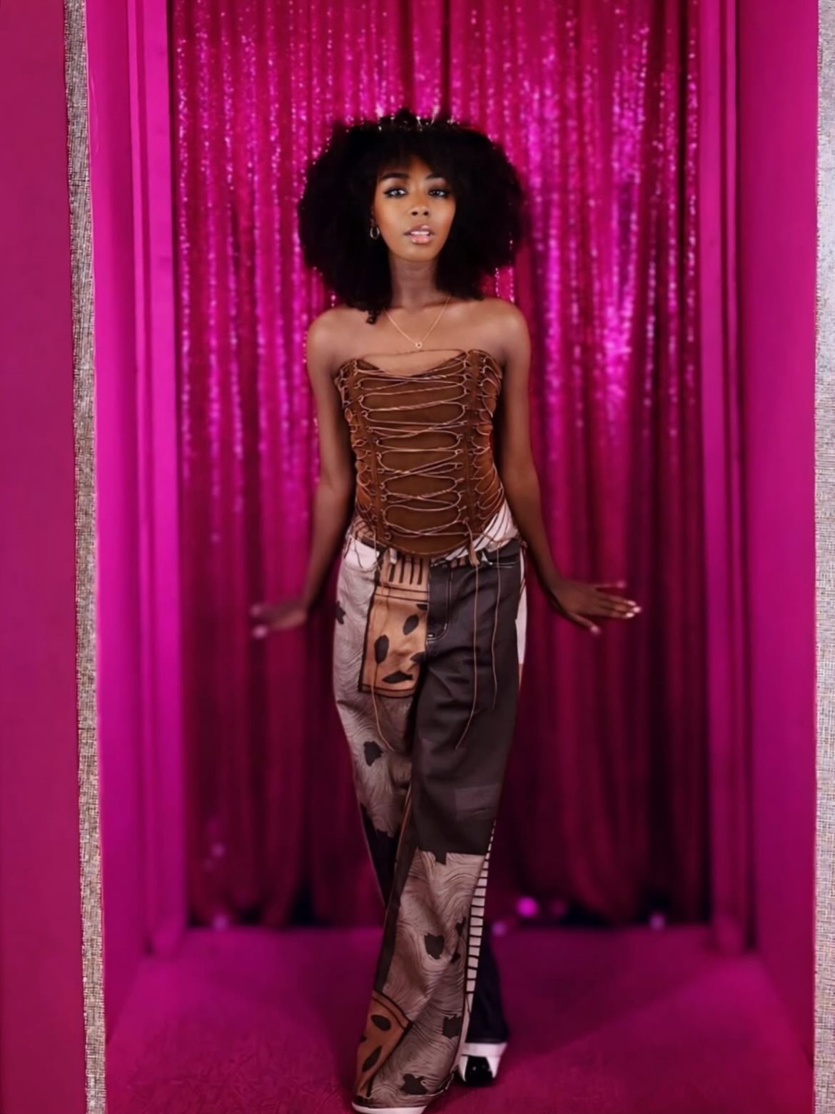 Young woman with curly hair stands in front of pink curtain, wearing a strapless brown top and patterned pants.