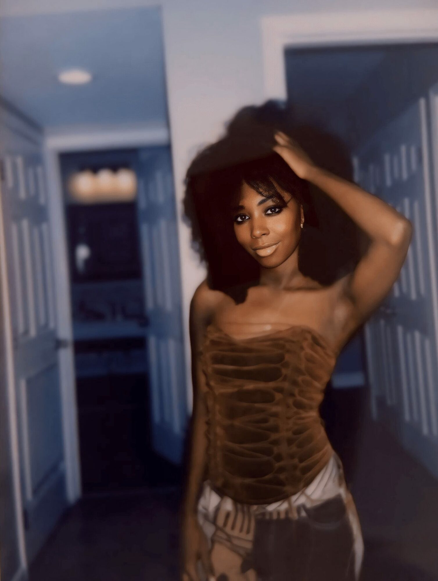 Young woman with curly hair in a hallway, wearing a strapless top and patterned pants, smiling and touching her hair.