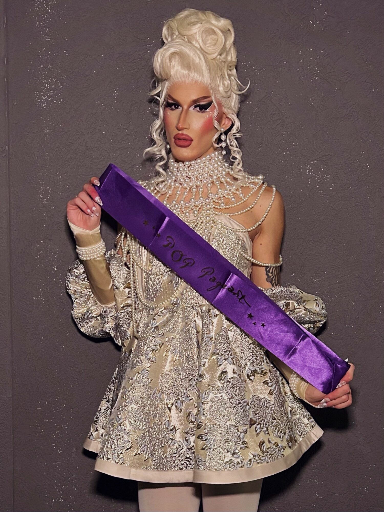 Person with elaborate hairstyle and makeup holding a purple sash, wearing a detailed silver dress with jewelry, against a plain background.