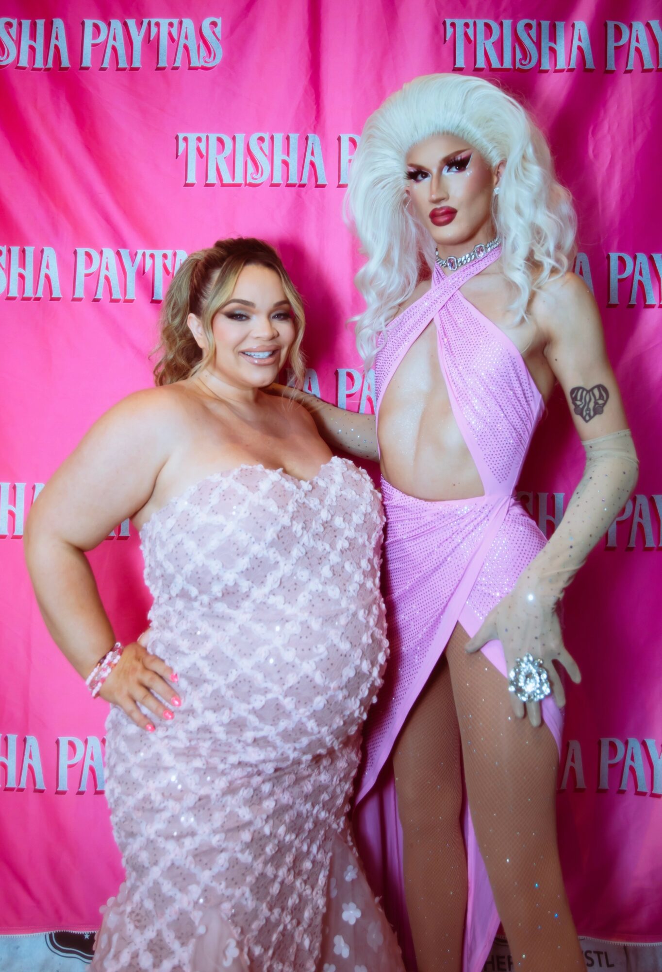 Two women pose in front of a pink backdrop with 'Trisha Paytas' written repeatedly. One woman is in a pink dress, the other in a patterned outfit.