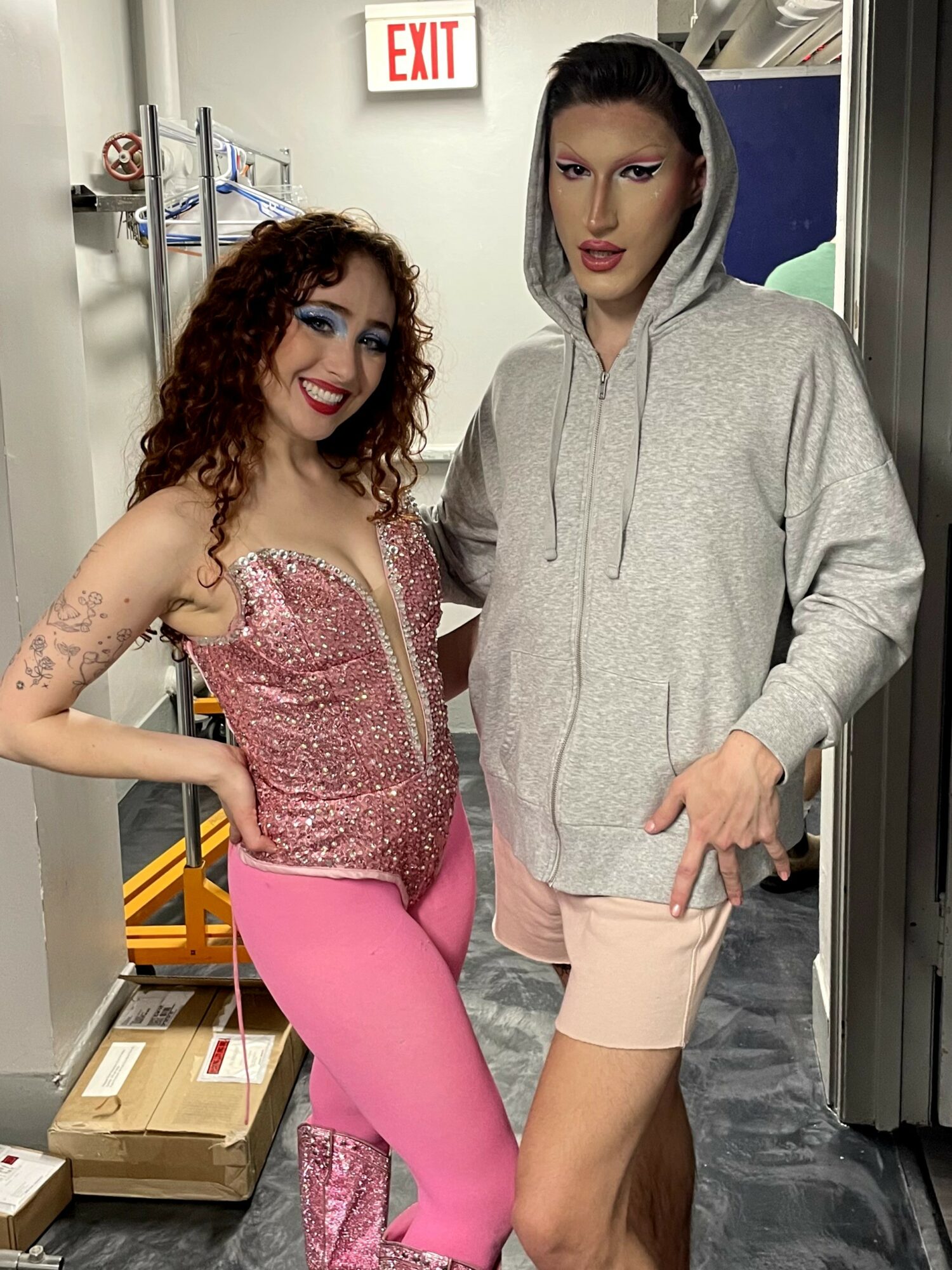 Two people standing indoors, one smiling with curly hair and pink outfit, the other in a gray hoodie and shorts.