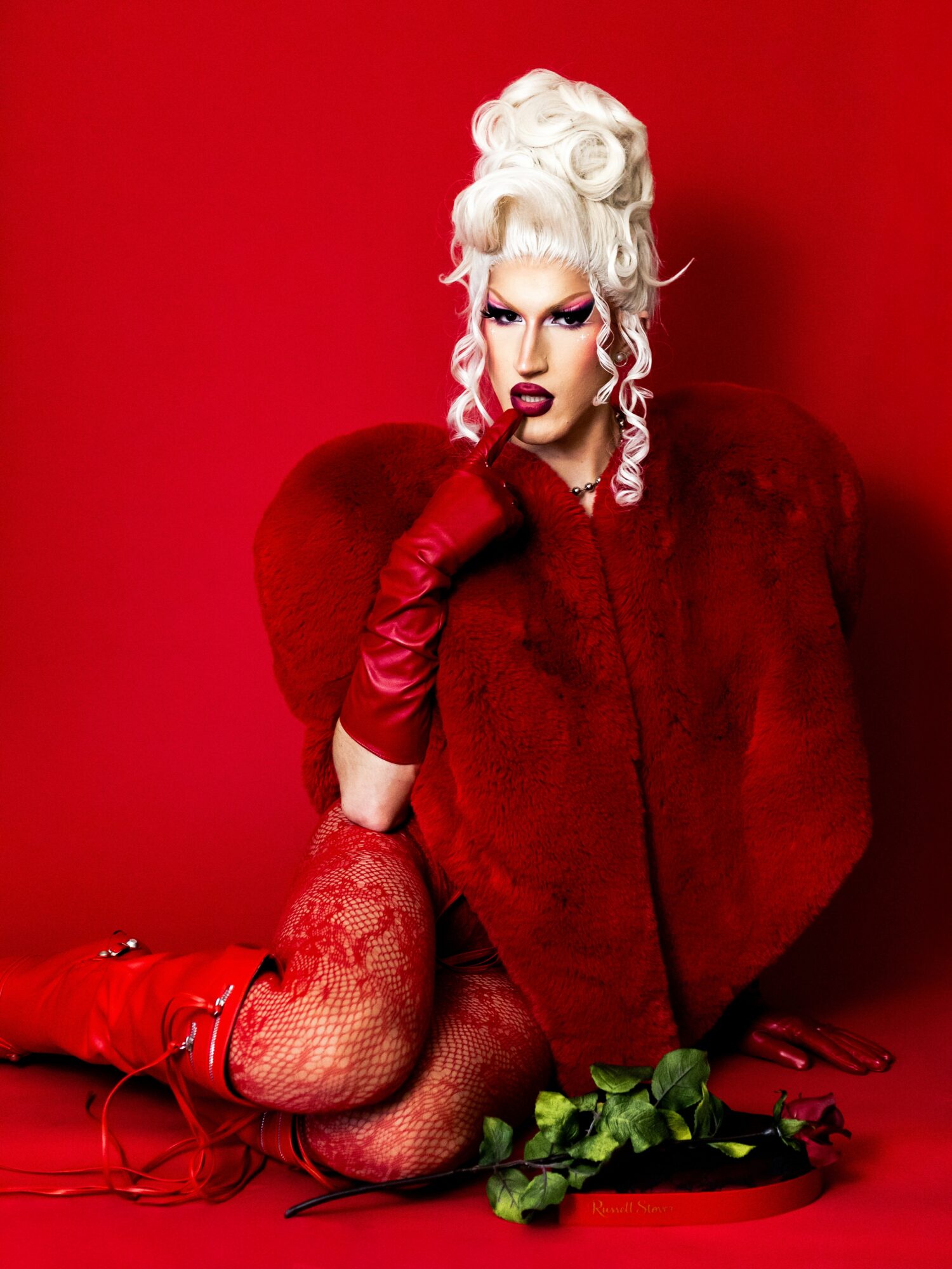 Person with elaborate white hairstyle, red fur coat, red gloves, and fishnet stockings, sitting against a red background.
