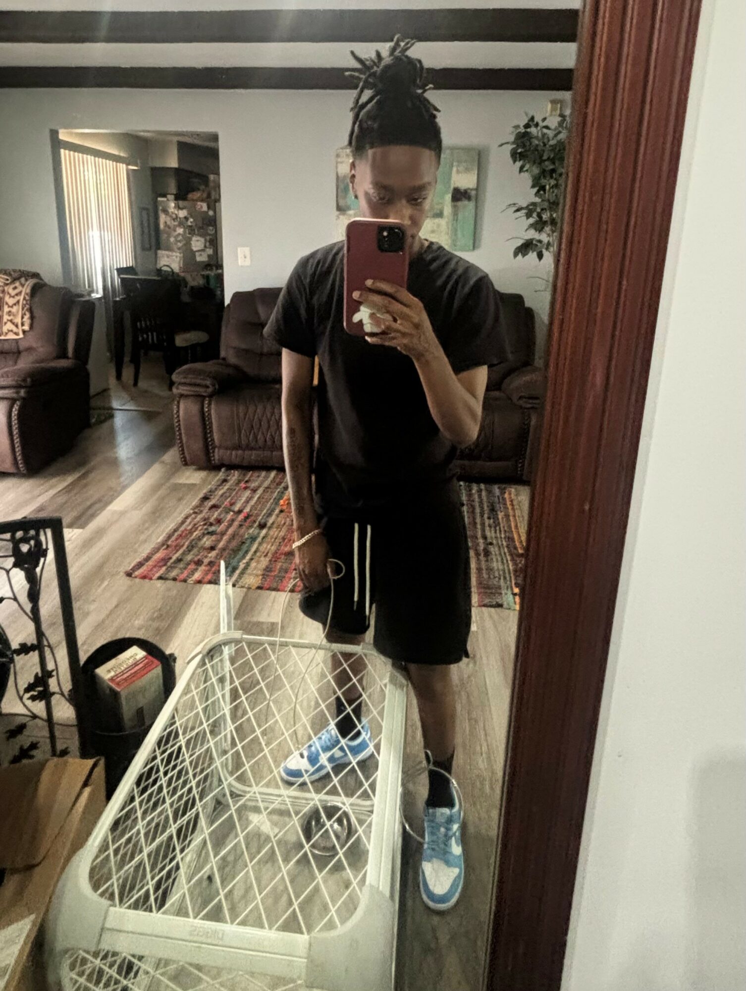 Person with dreadlocks taking a mirror selfie in a living room, wearing black shirt, shorts, and sneakers, holding a phone.