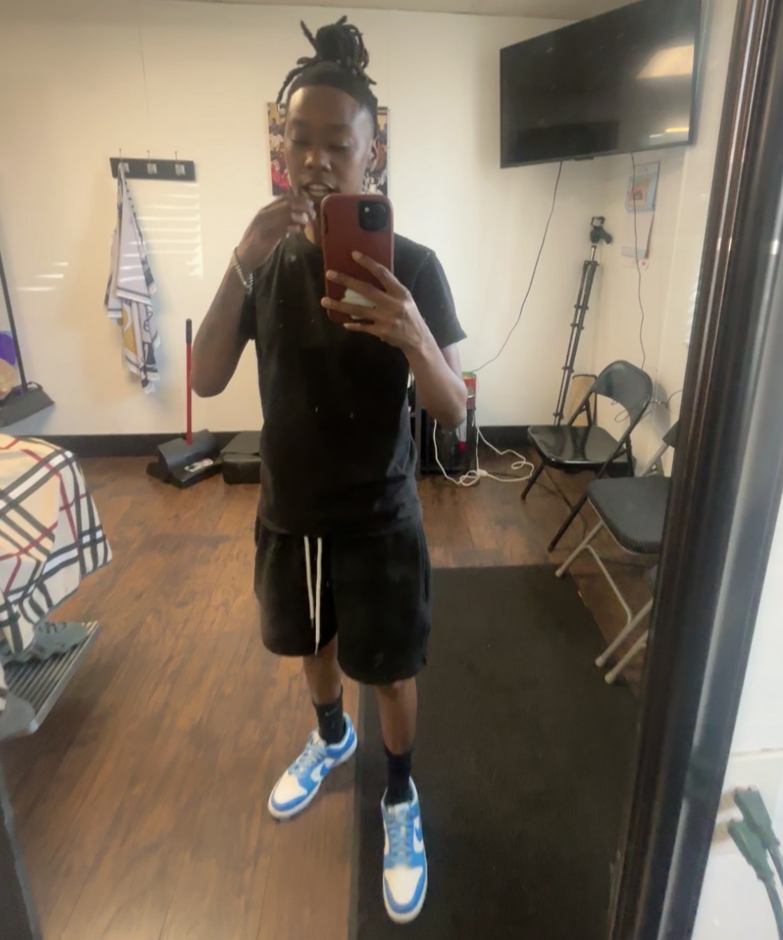 Person taking a mirror selfie in a room with a TV, chairs, and a towel rack, wearing black shirt, shorts, and blue sneakers.