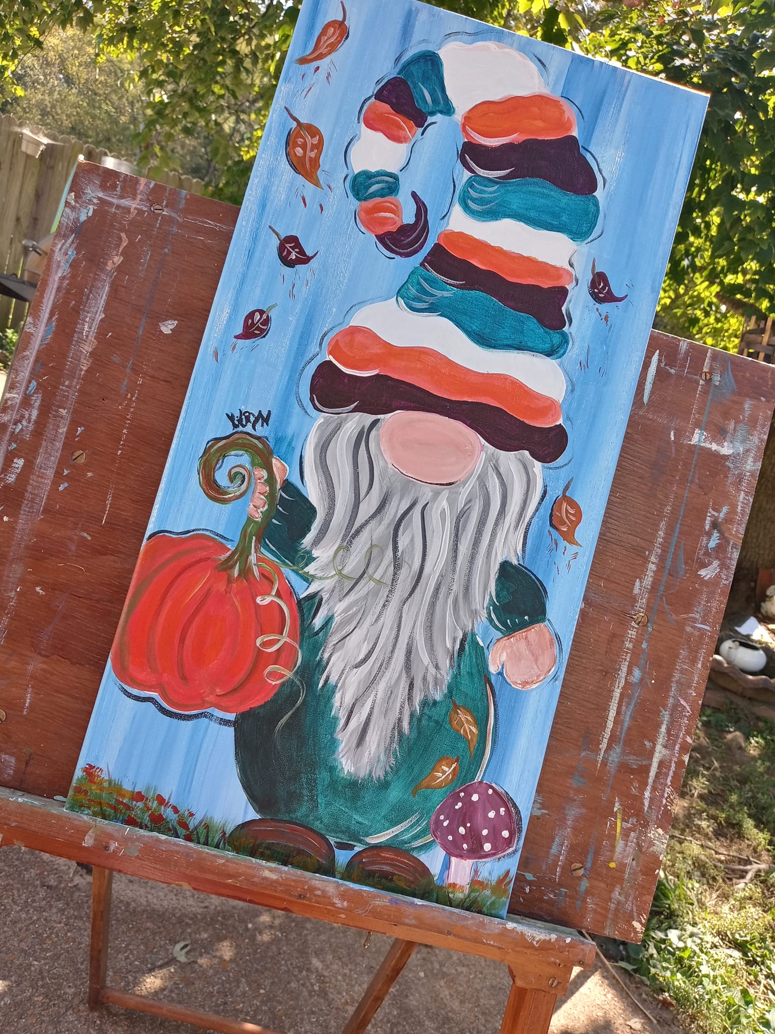 Painting of a gnome with a tall, colorful striped hat, long white beard, holding a pumpkin, surrounded by mushrooms and butterflies.