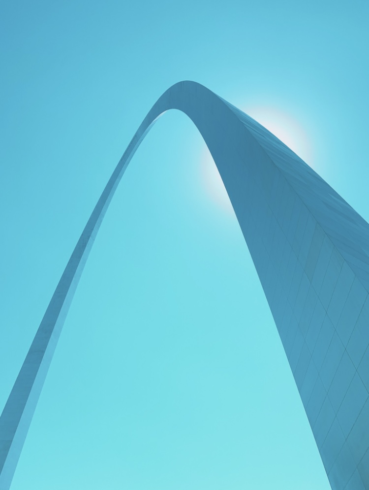 Looking up at a tall, curved skyscraper against a bright blue sky with the sun shining behind it.