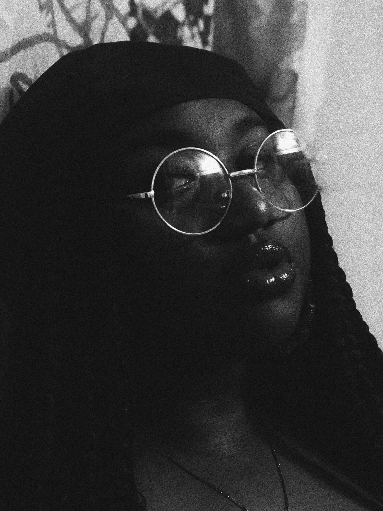 Person wearing round glasses and a head covering, with braided hair, in black and white.