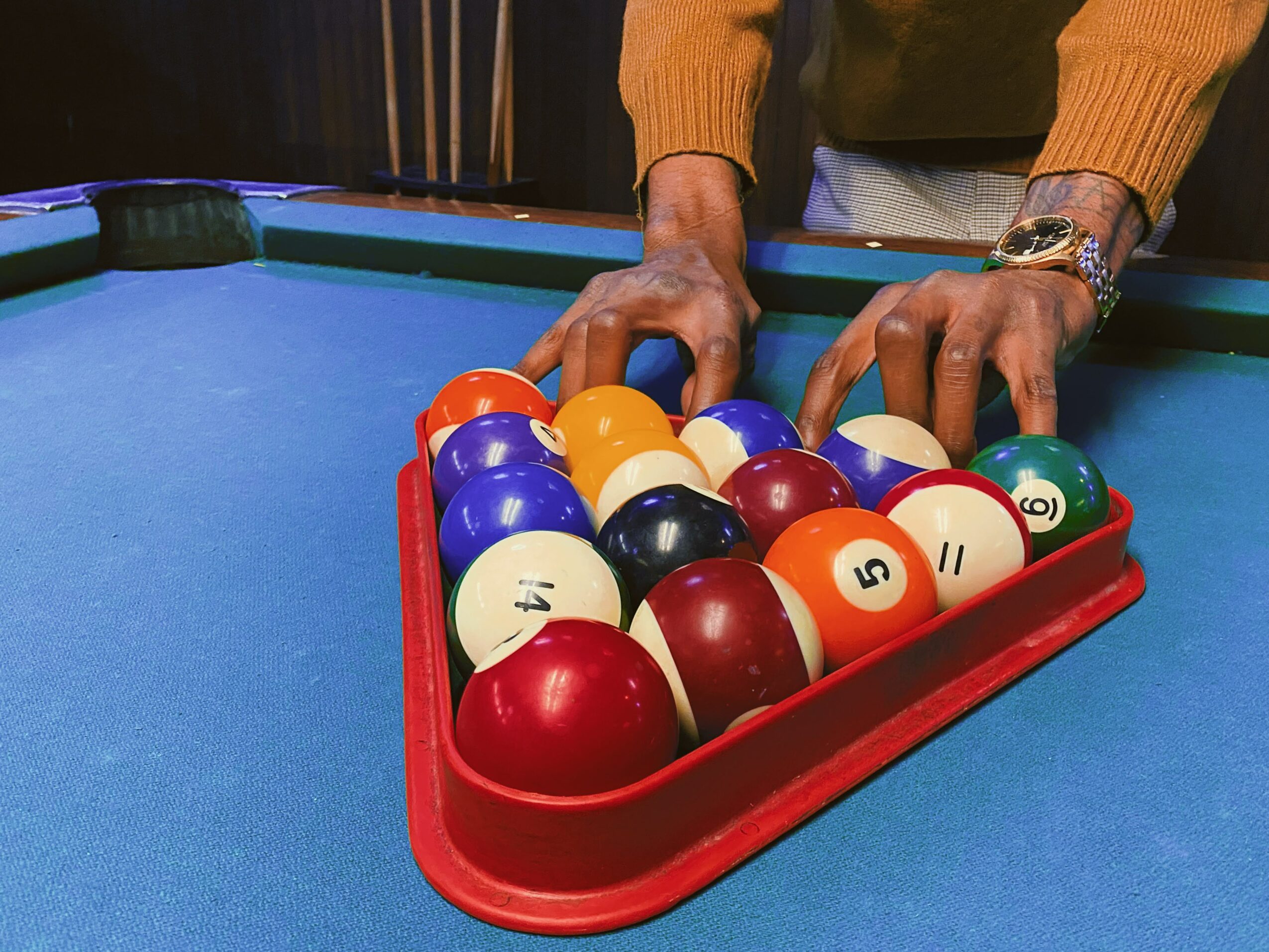 Person's hands reaching for billiard balls on a pool table with a red tray, blue felt surface, and a watch on the wrist