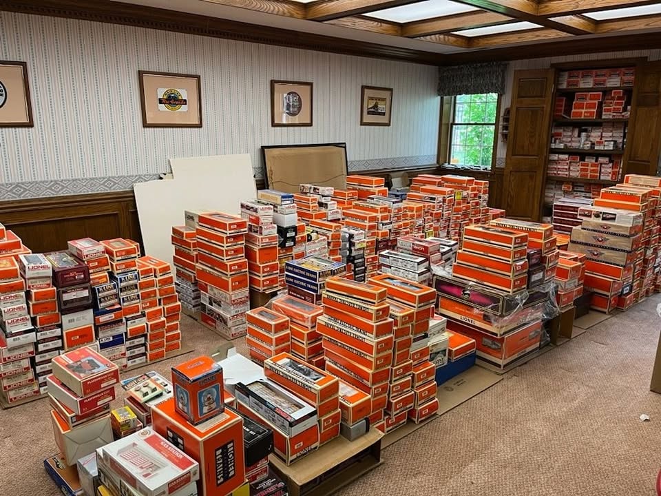 Room filled with stacks of books on tables and shelves, with framed pictures on the wall and a window showing greenery.