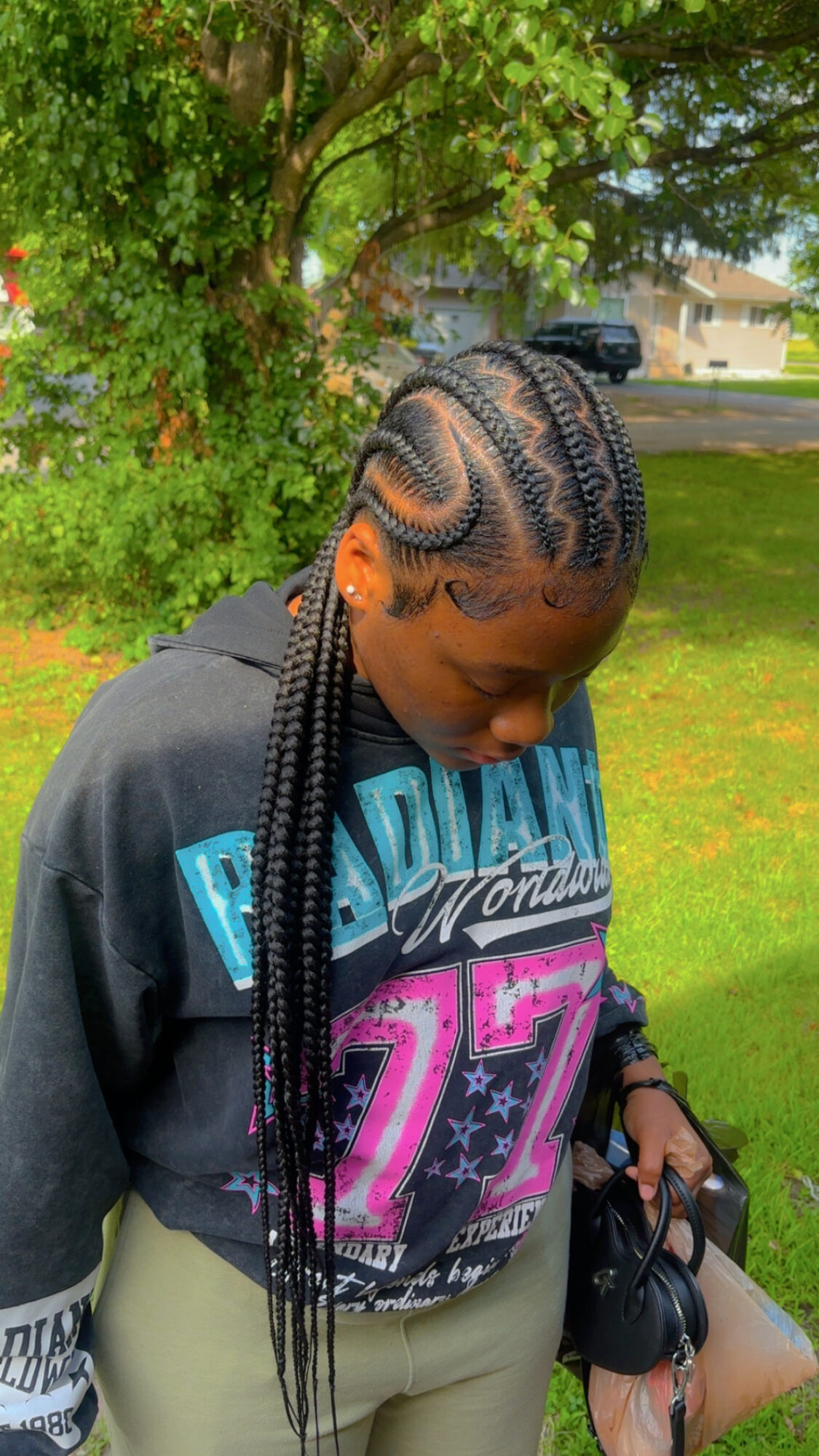 Young woman with braided hair outdoors, wearing a black hoodie with colorful text, holding headphones, standing on grass near trees.