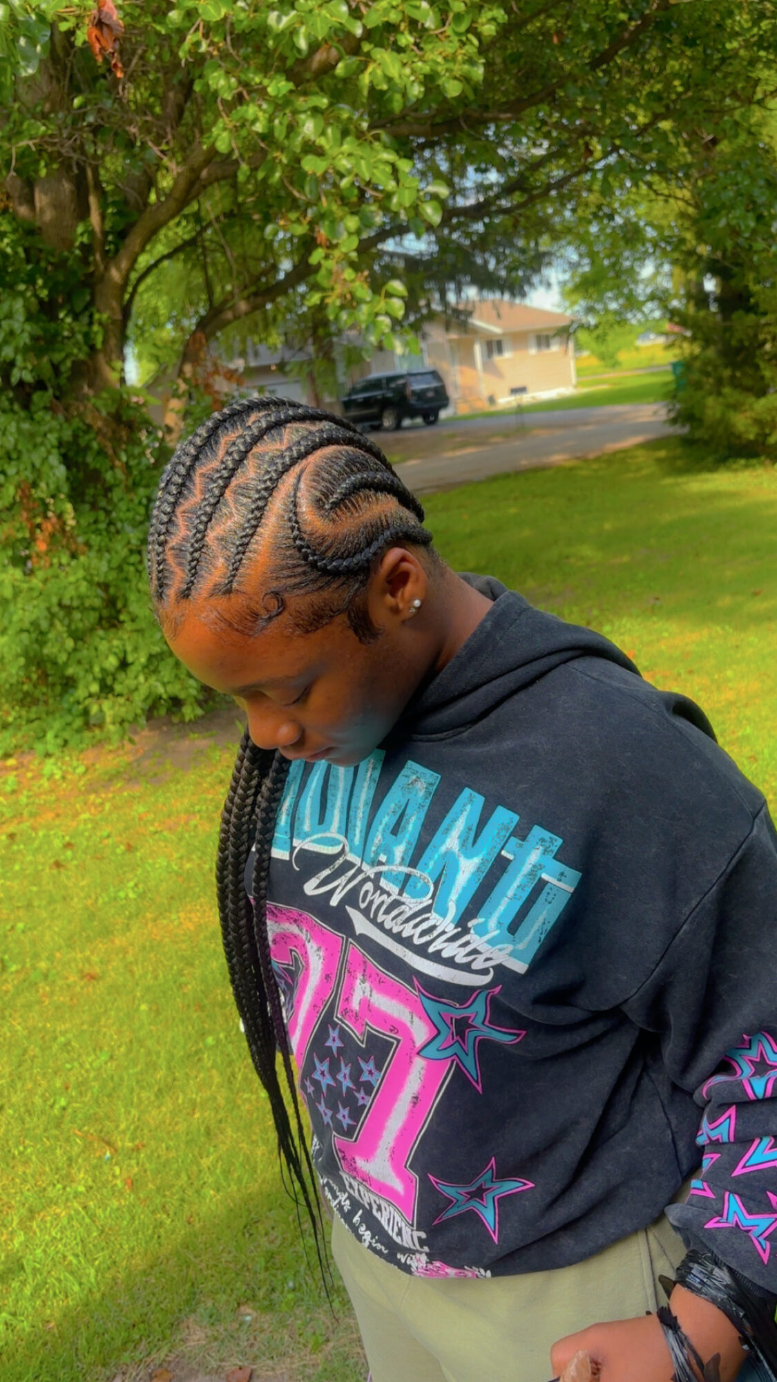 Young person with braided hair looking down outdoors near trees and grass.