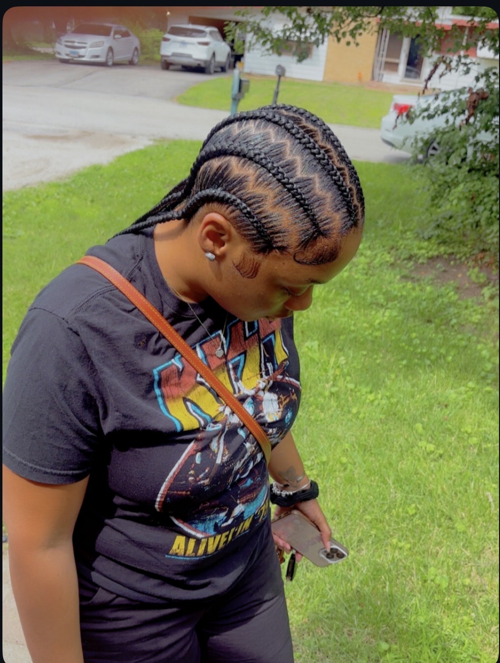 Young woman with braided hair looking down at her phone outdoors on grass, with cars and buildings in background.