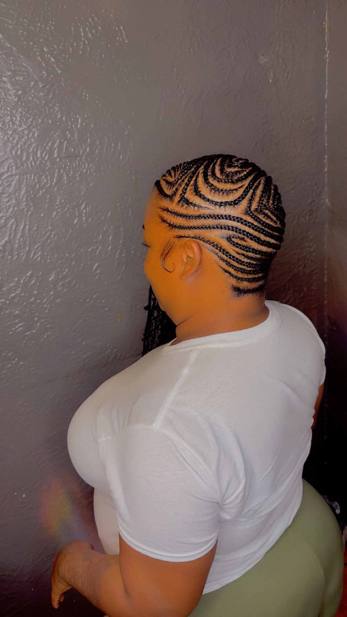 Person with intricate braided hairstyle facing a gray wall, wearing a white shirt and light-colored pants.
