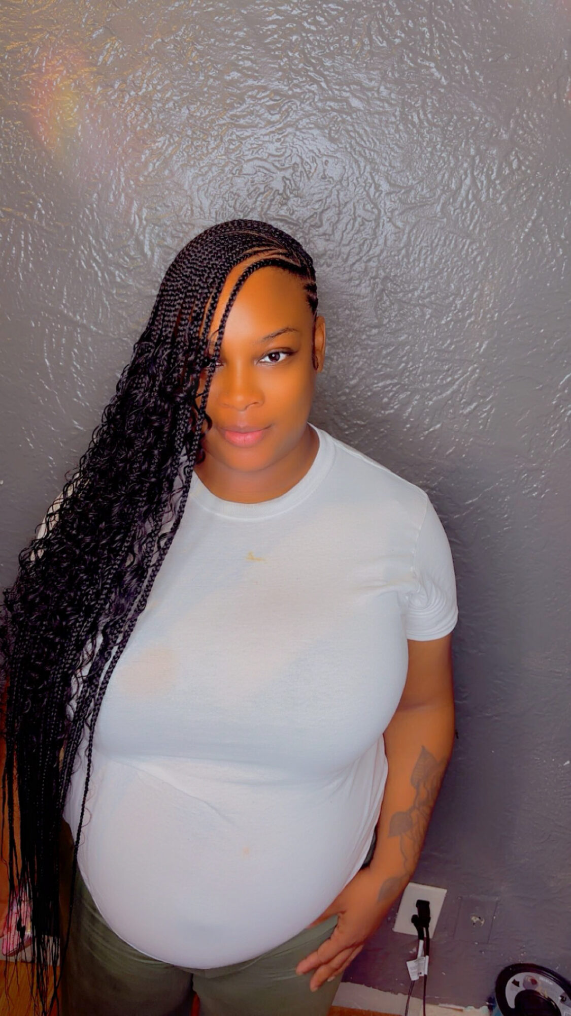 Woman with long braided hair wearing a white shirt, standing against a gray textured wall.