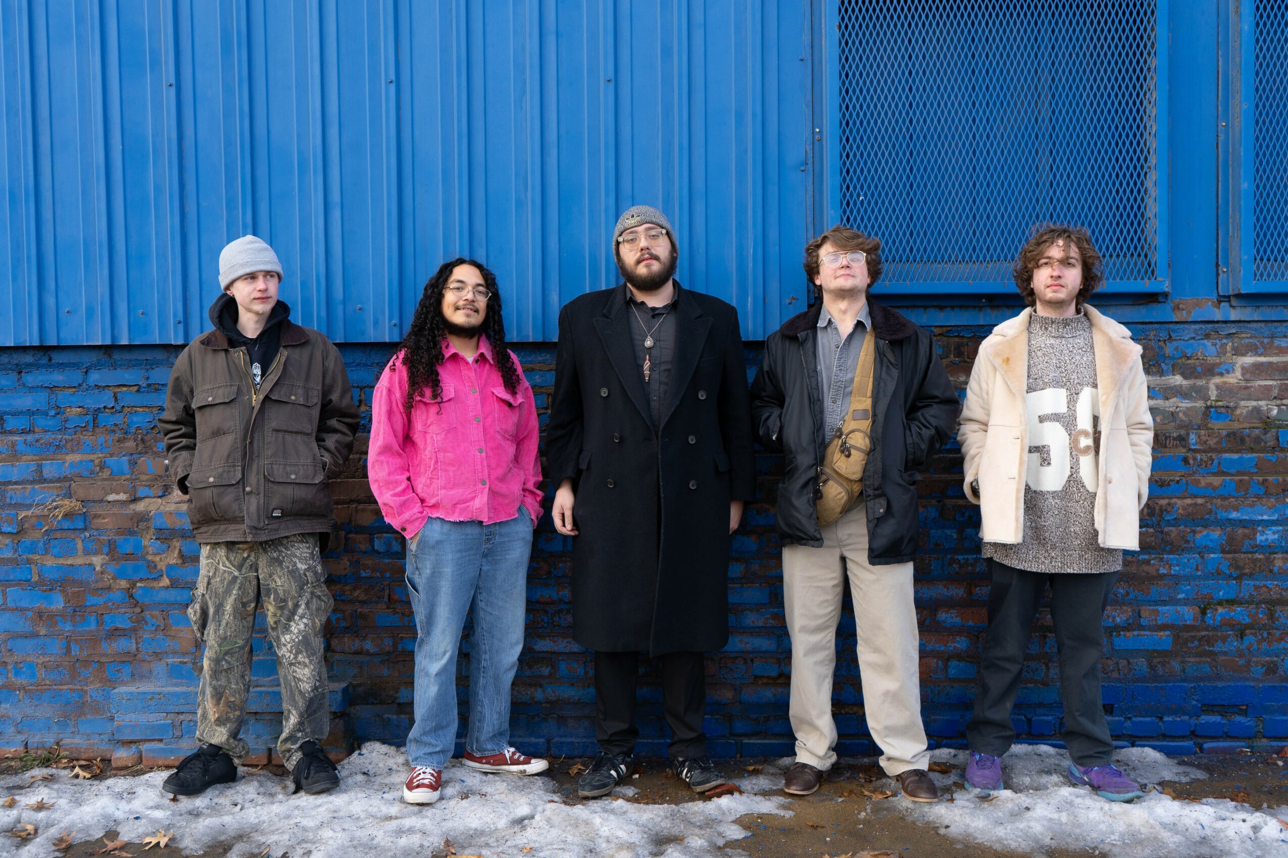 Five people stand outdoors in front of a blue wall and brick wall, dressed in casual clothing, some with jackets and hats.