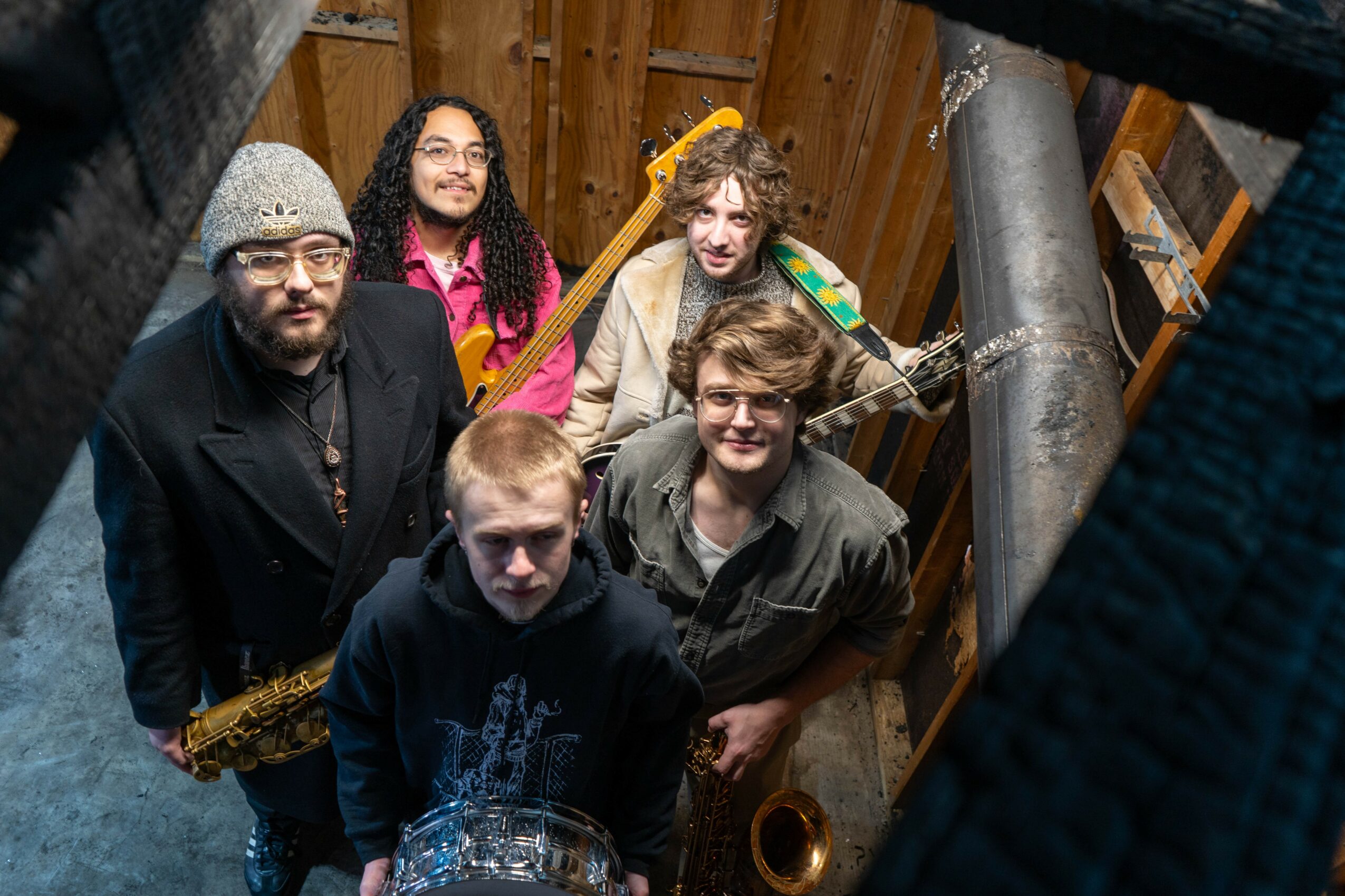 Group of five people in a wooden room, some holding musical instruments, looking up at the camera from above.