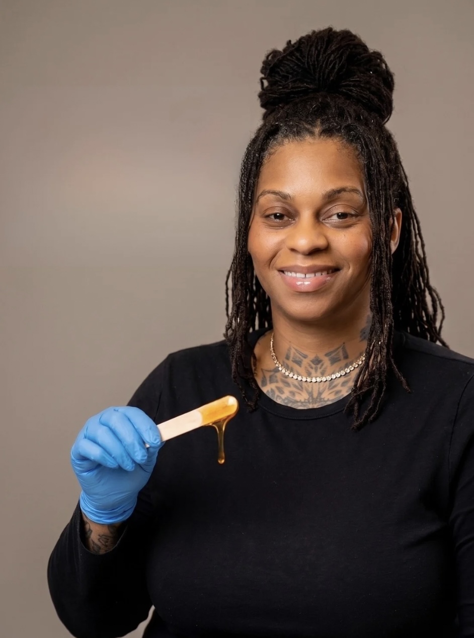 Woman with braided hair in a bun, wearing a black top and blue glove, holding a honey dipper with honey, smiling.