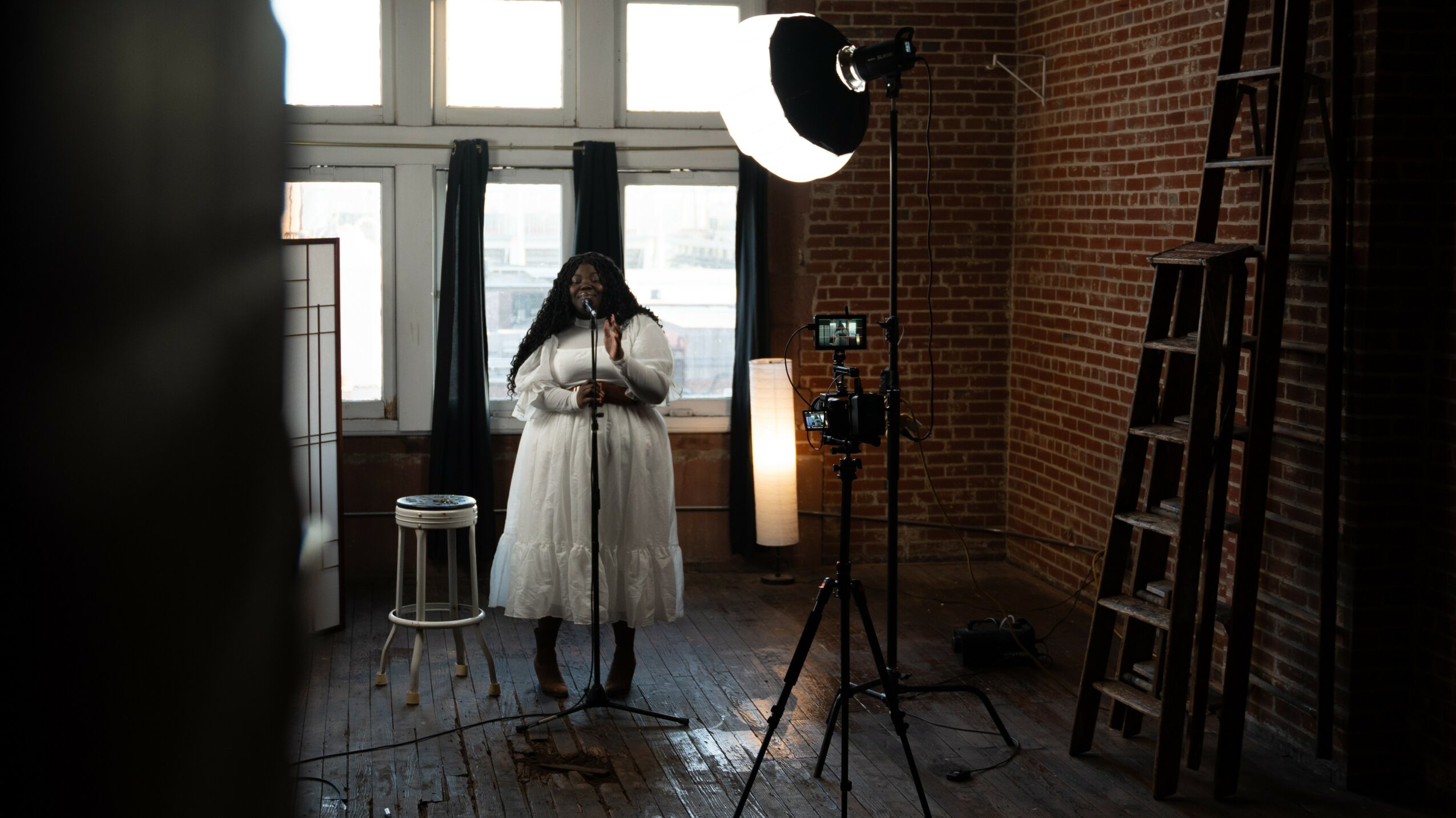 Person in white dress standing in a room with brick walls, large windows, and photography equipment, facing forward.