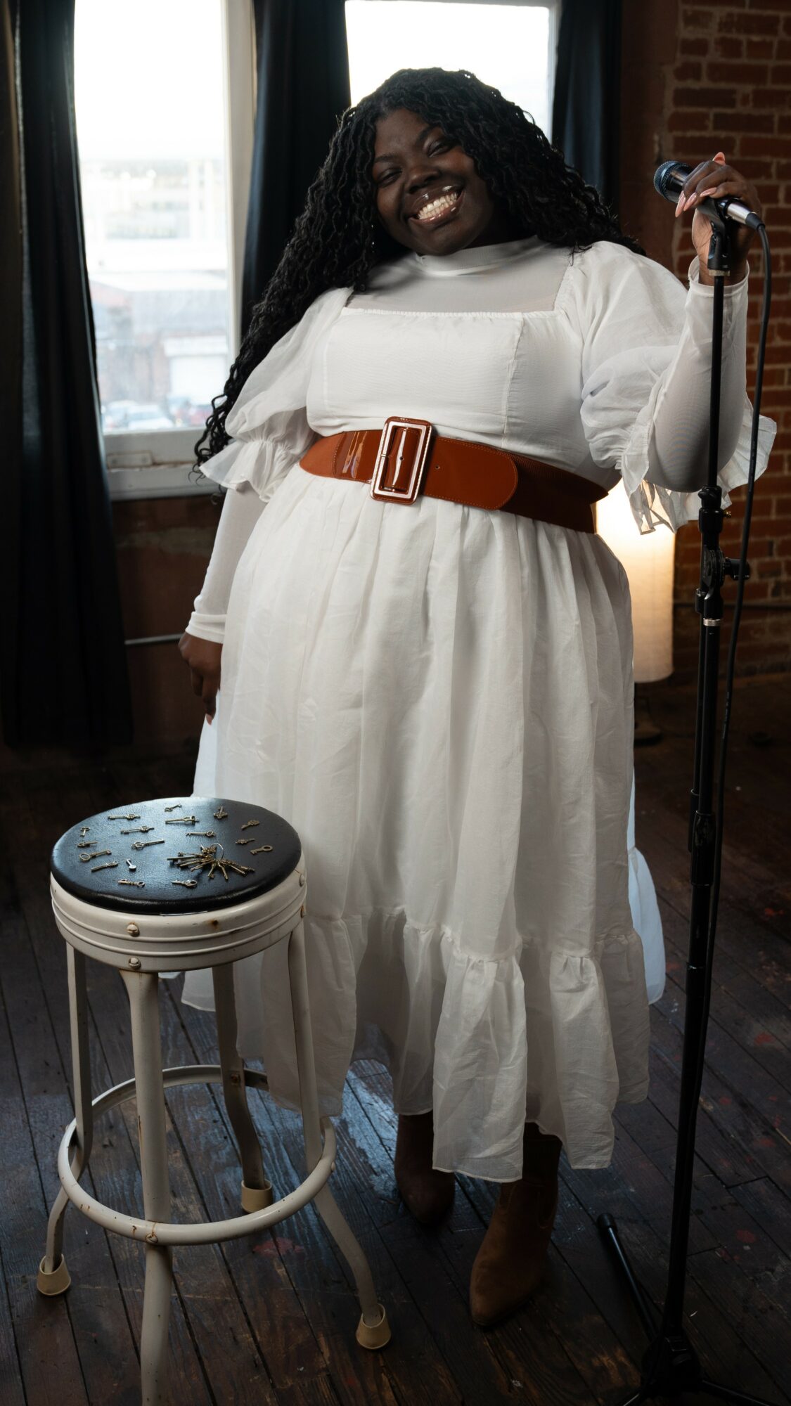 Person with long curly hair in a white dress with a wide belt, smiling, standing near a microphone and a stool.