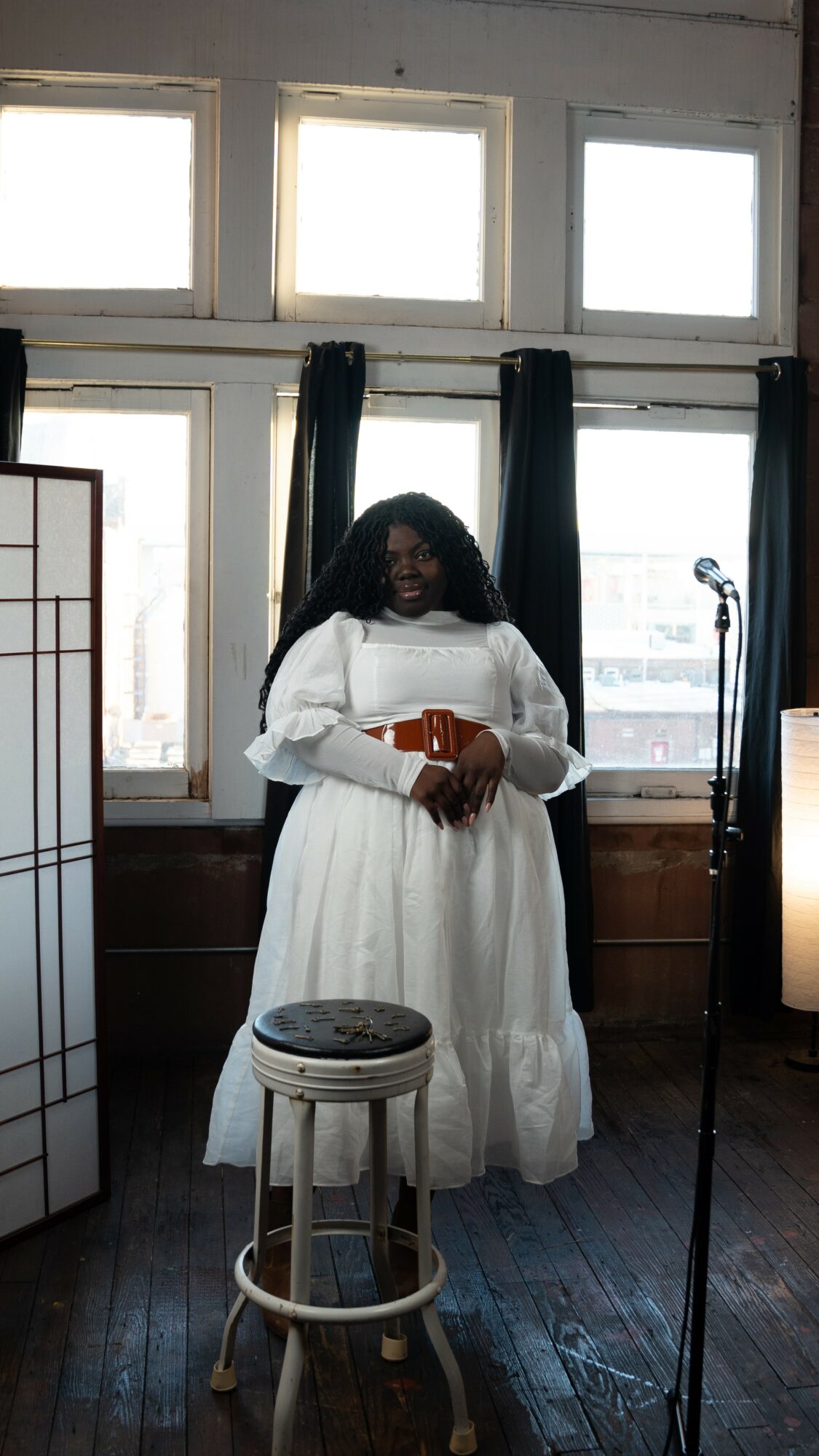Woman in white dress holding a book, standing in front of windows with black curtains, in a room with wooden floors.
