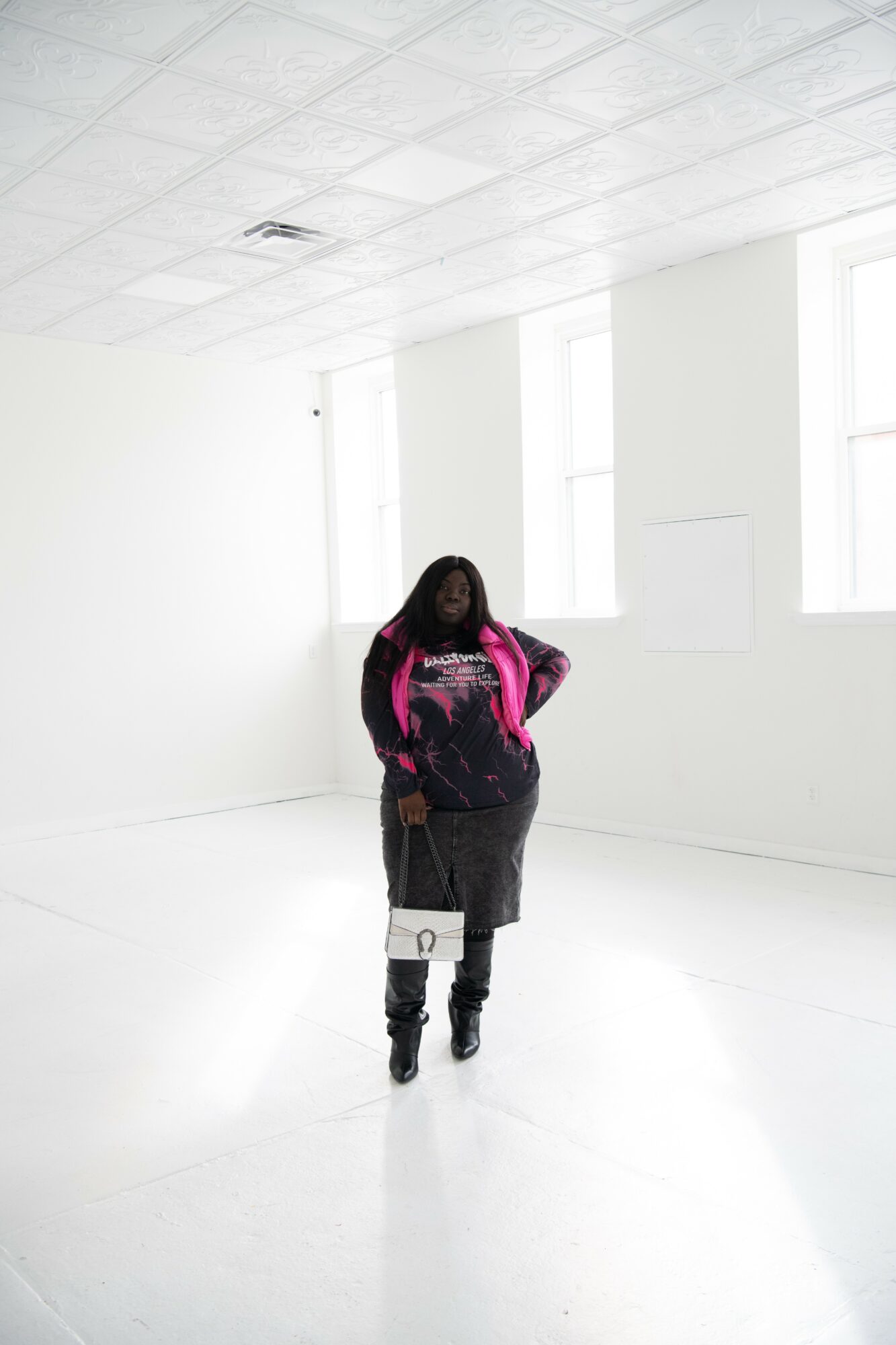 Person standing in a bright, white room with large windows, wearing a black dress and pink jacket, hands on hips.