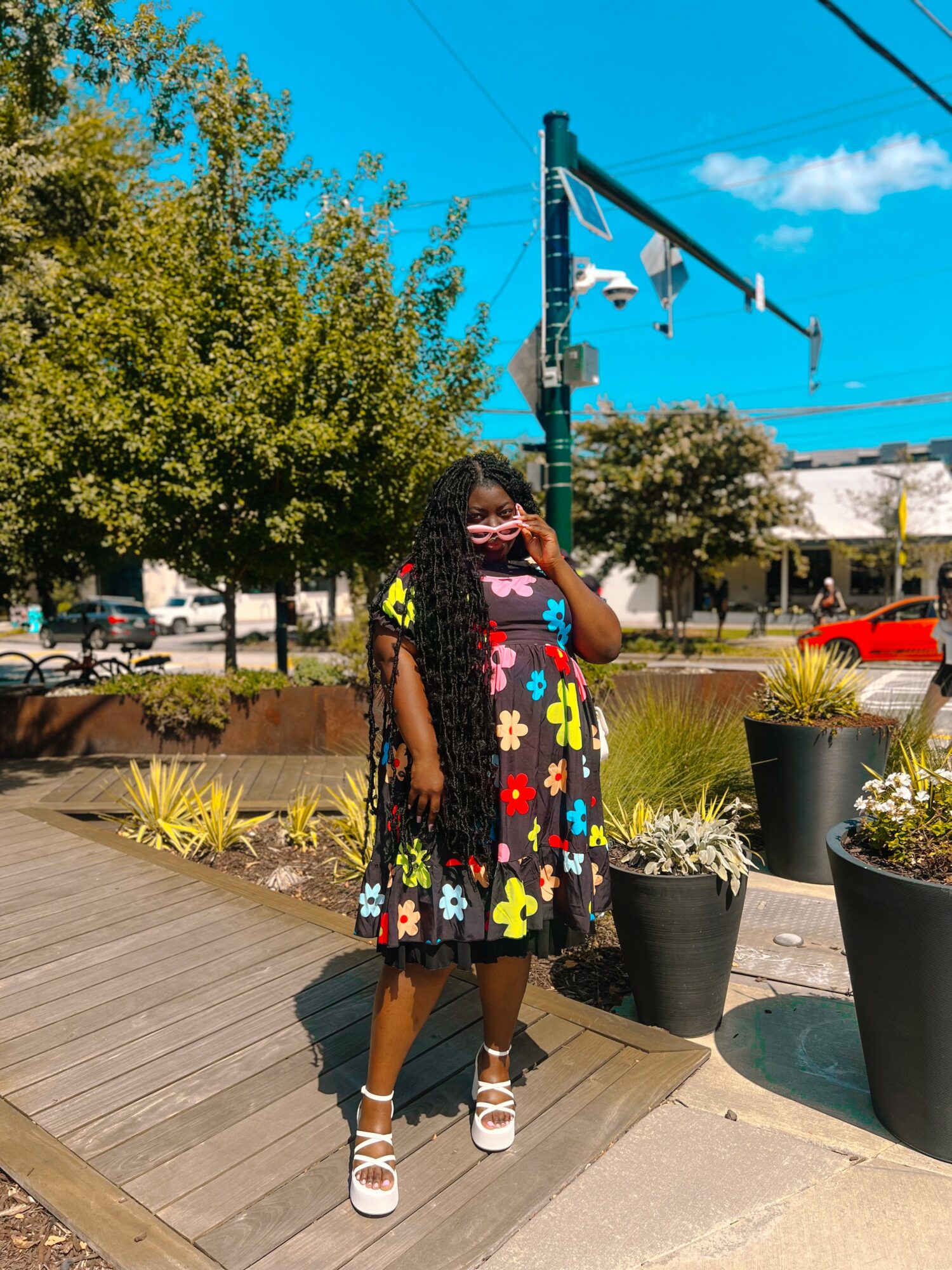 Person wearing a colorful dress with floral patterns, standing outdoors near potted plants and trees, talking on a phone.