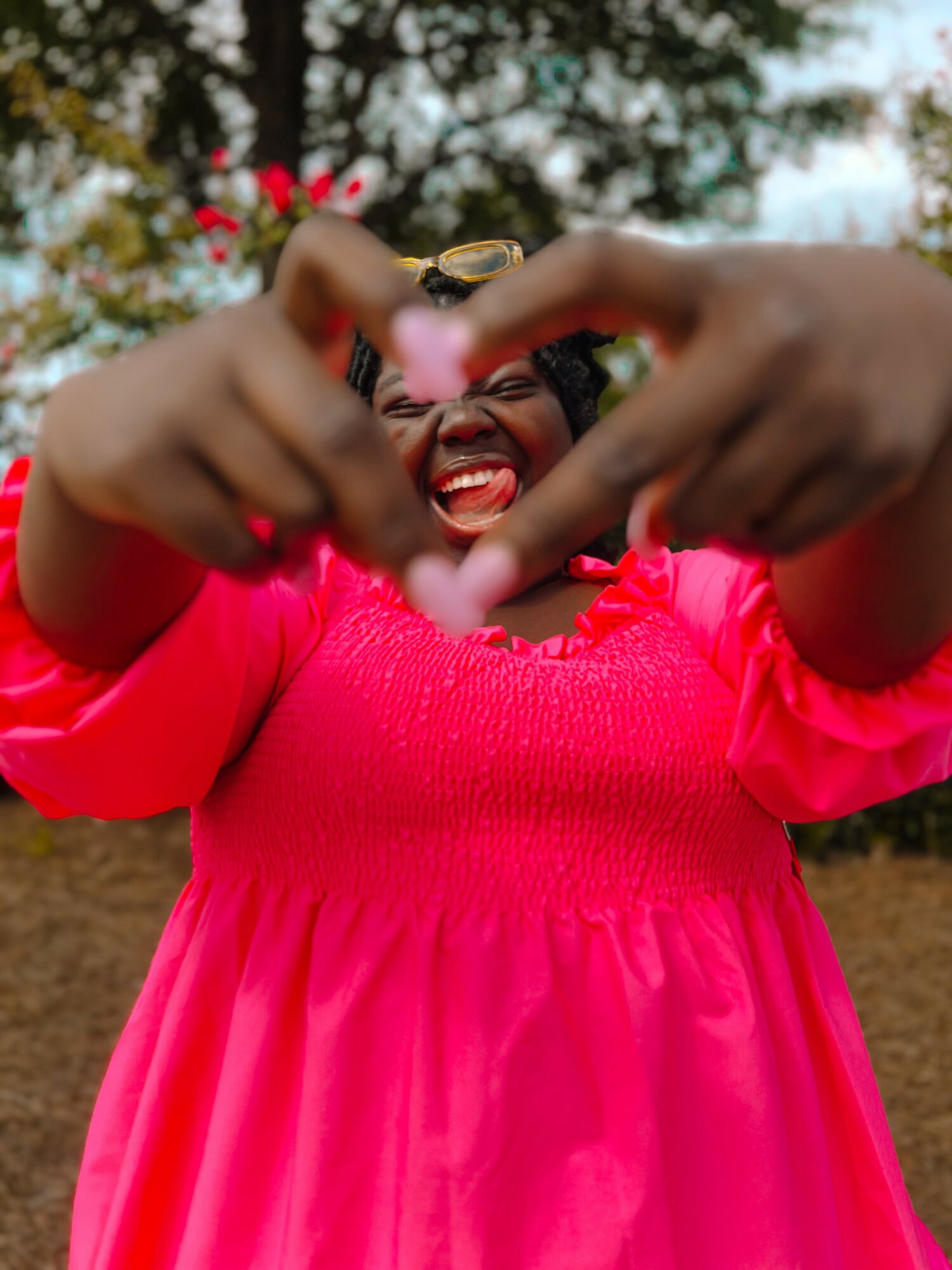 Woman smiling outdoors, forming a heart shape with her hands, wearing a bright pink dress, trees in background.