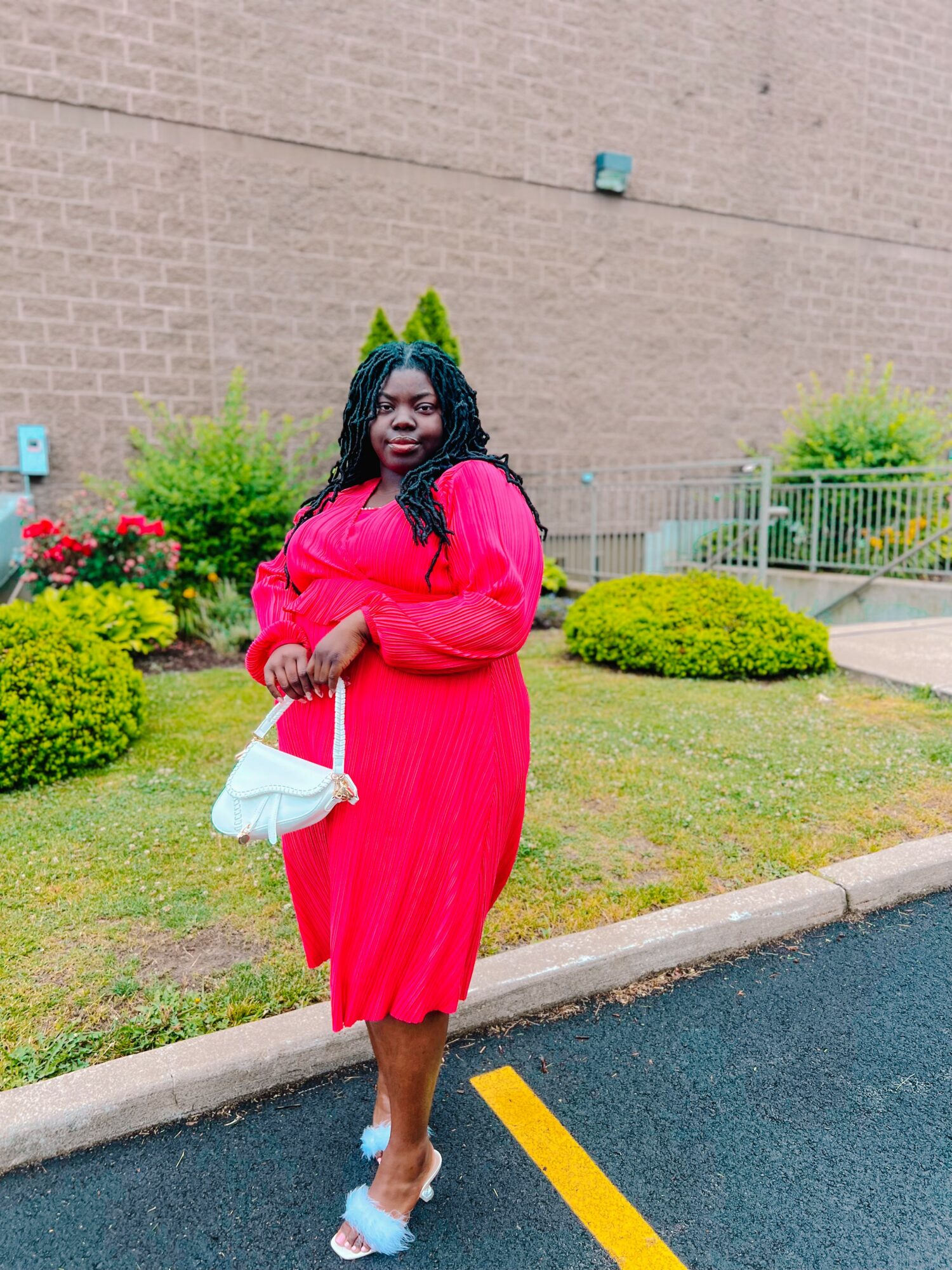 Woman in a bright pink dress holding a white purse, standing outdoors near a brick wall and greenery.