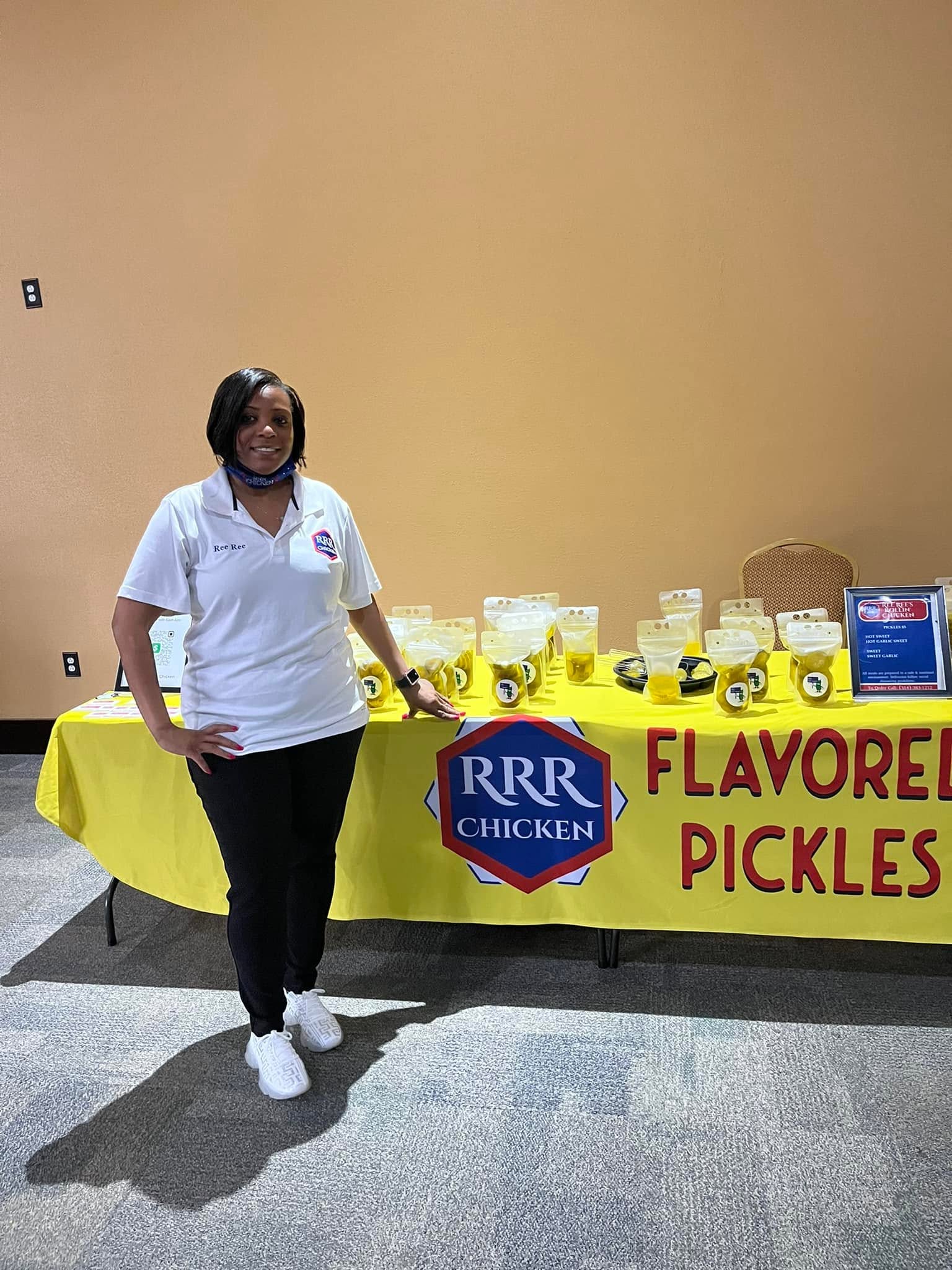 Woman standing next to a table with jars of pickles and a sign that reads 'Flavored Pickles'.