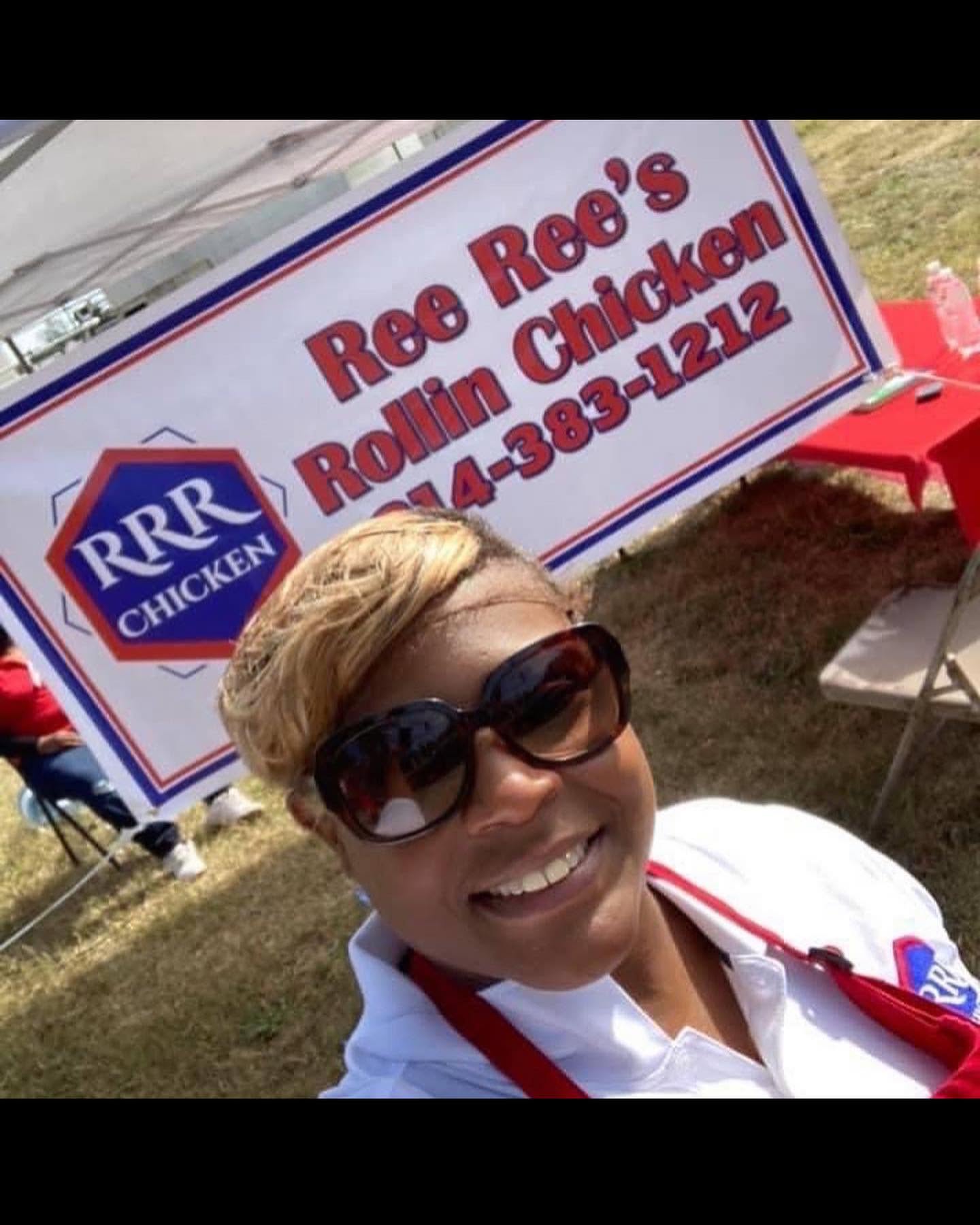 Woman with short hair and sunglasses smiling outdoors near a sign for Ree Ree's Rollin Chicken, with red chairs nearby.