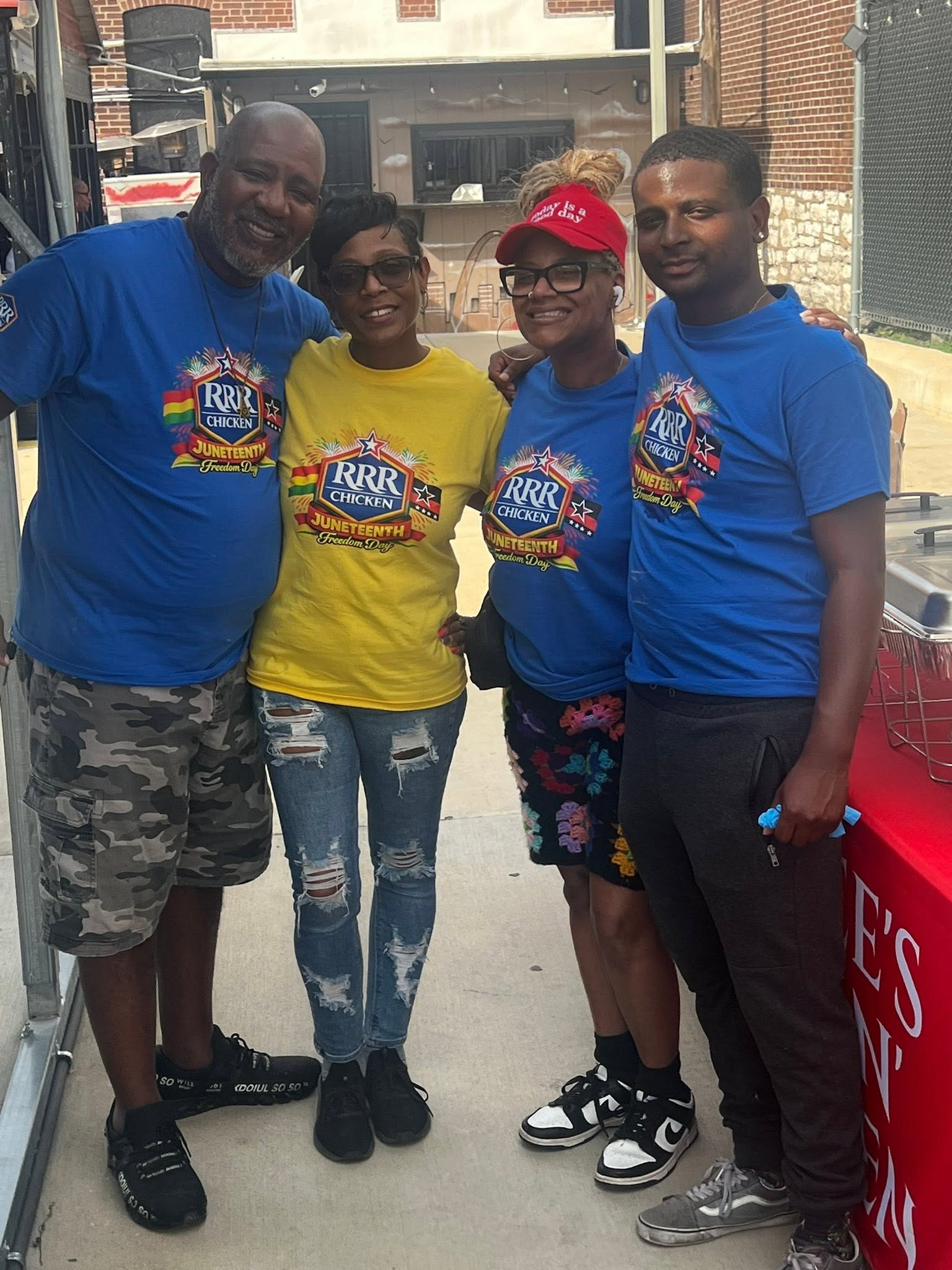 Four people standing outdoors, smiling, wearing colorful shirts, with buildings and a red object in the background.
