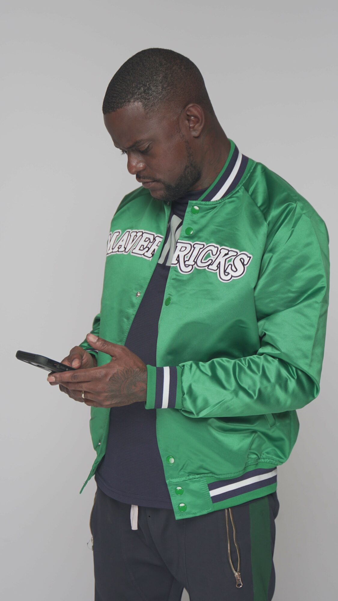 Man in green jacket looking at phone, standing sideways against a plain background.