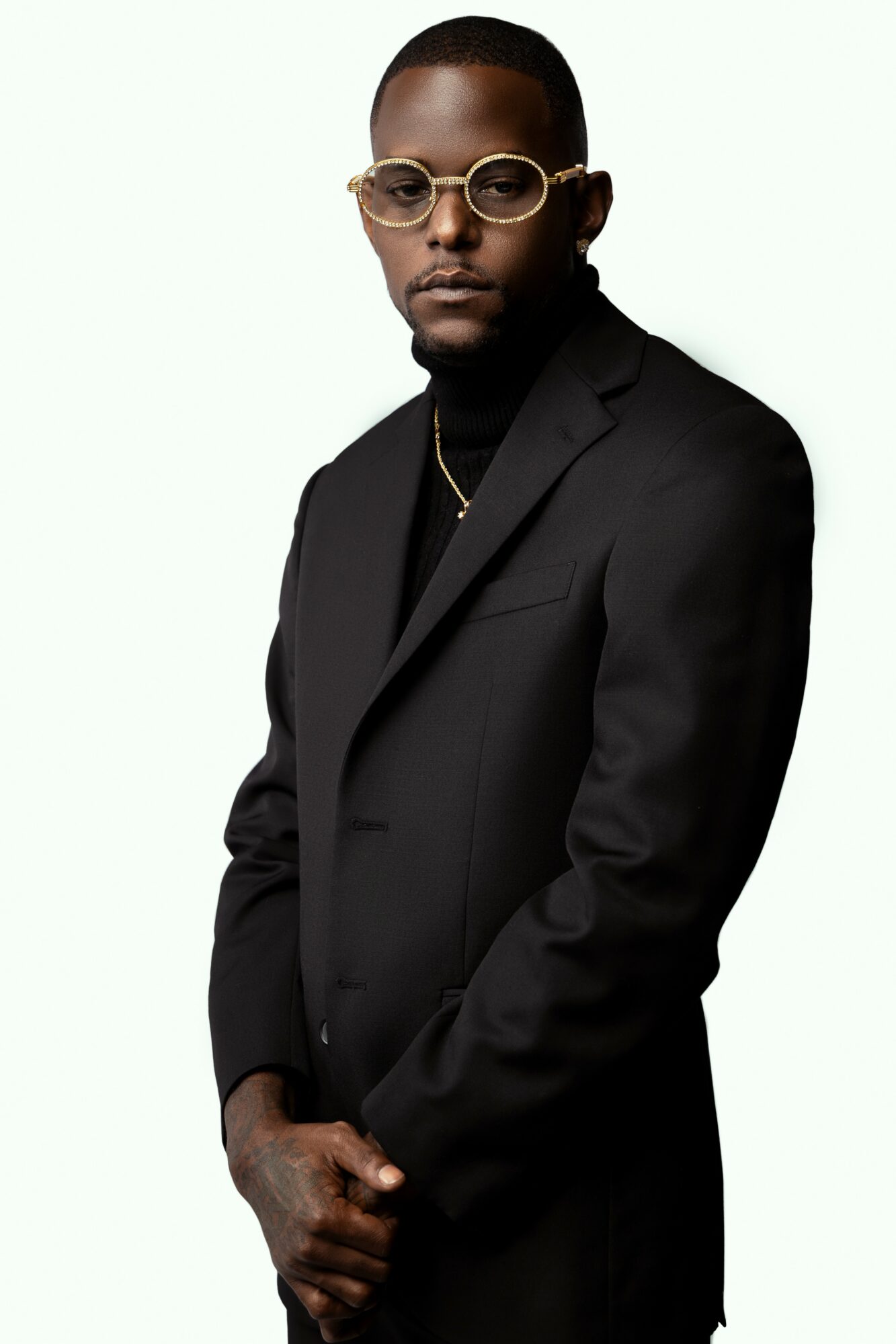 Man in black suit with glasses and gold chain, standing with hands clasped, against a light background.