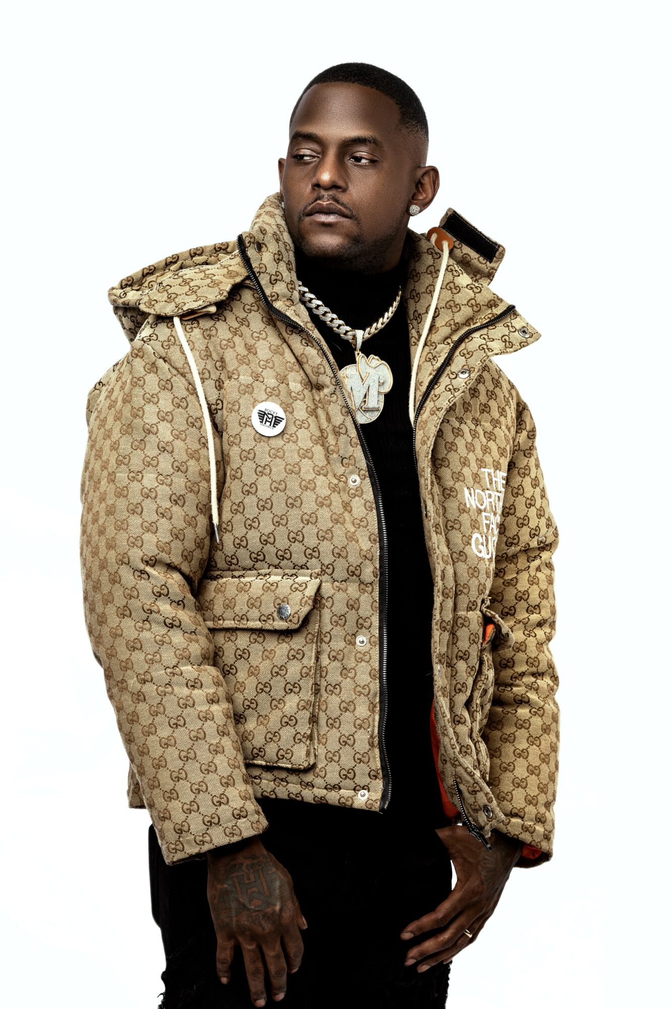 Man wearing a patterned beige jacket with a black shirt, jewelry, and a serious expression, standing against a white background.