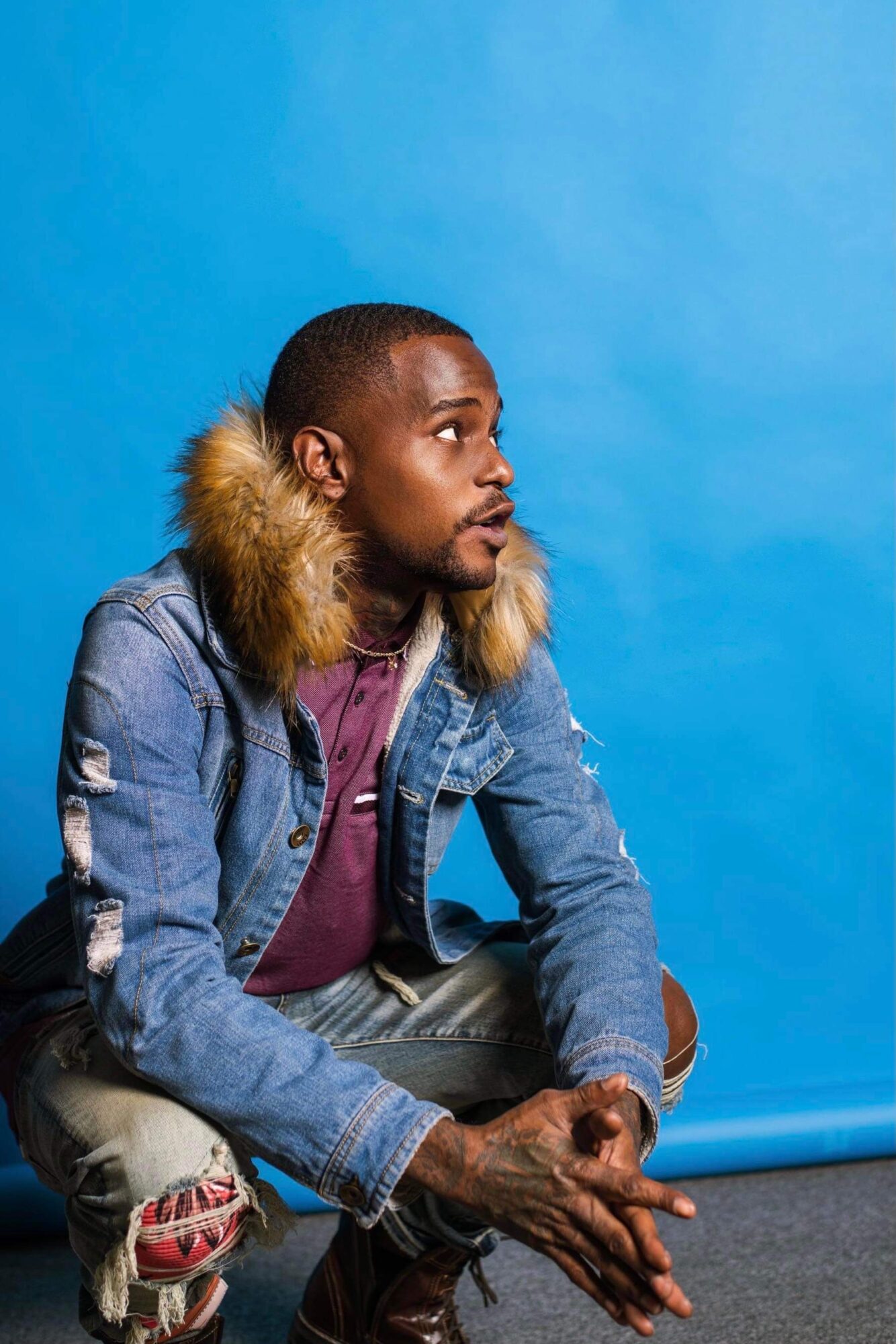 Young man with short hair and beard, wearing a denim jacket with fur collar, sitting against a blue background.