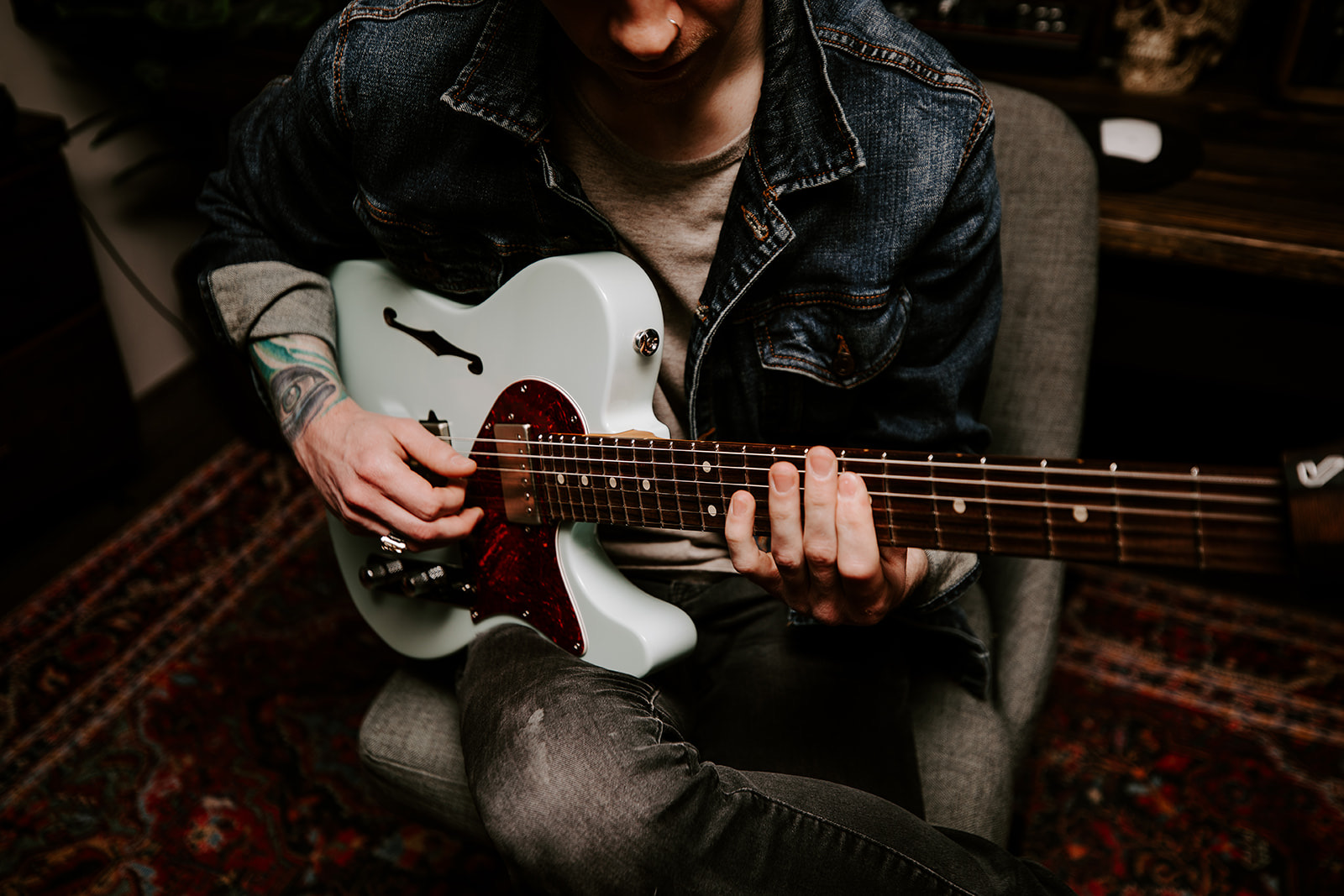 Person playing white electric guitar while seated, wearing a denim jacket, in a room with a patterned rug.