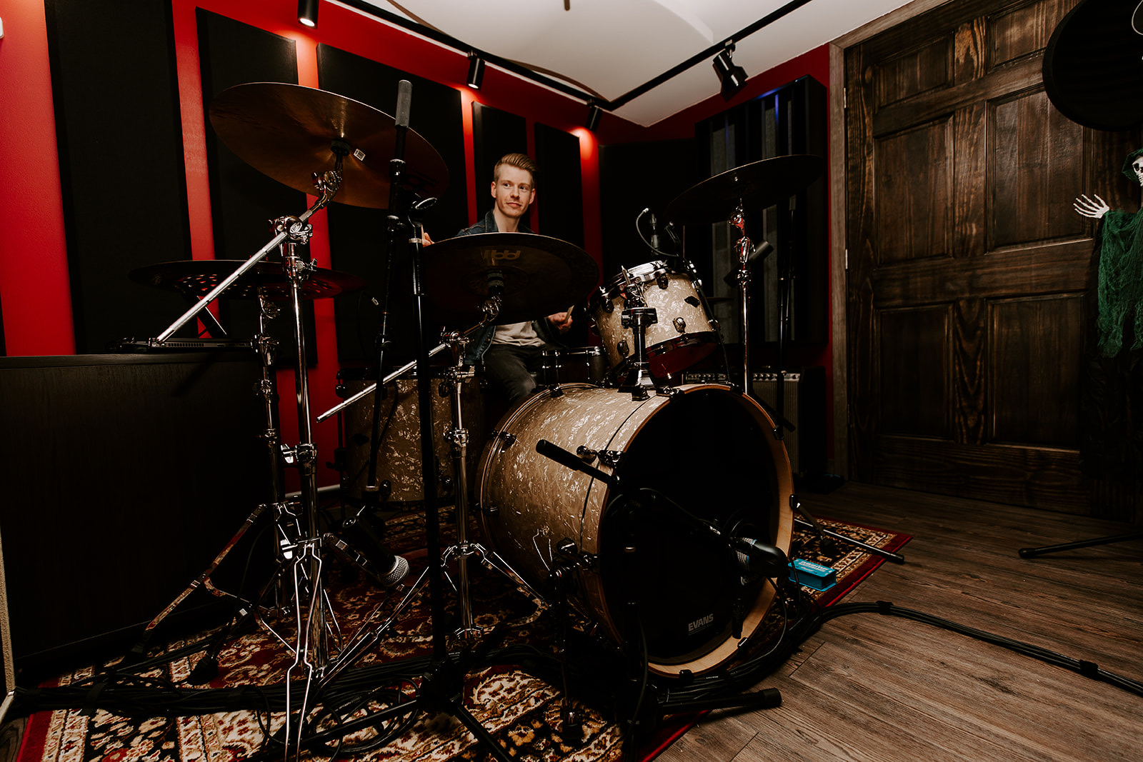 Person playing drums in a music studio with red and black walls, wooden door, and musical equipment.