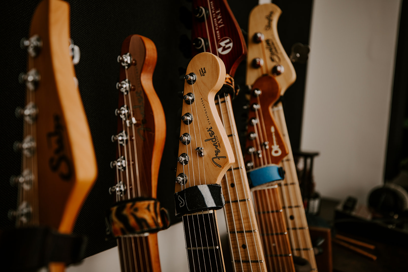 Four electric guitar headstocks hanging on a wall, with tuning pegs visible and a blurred background.