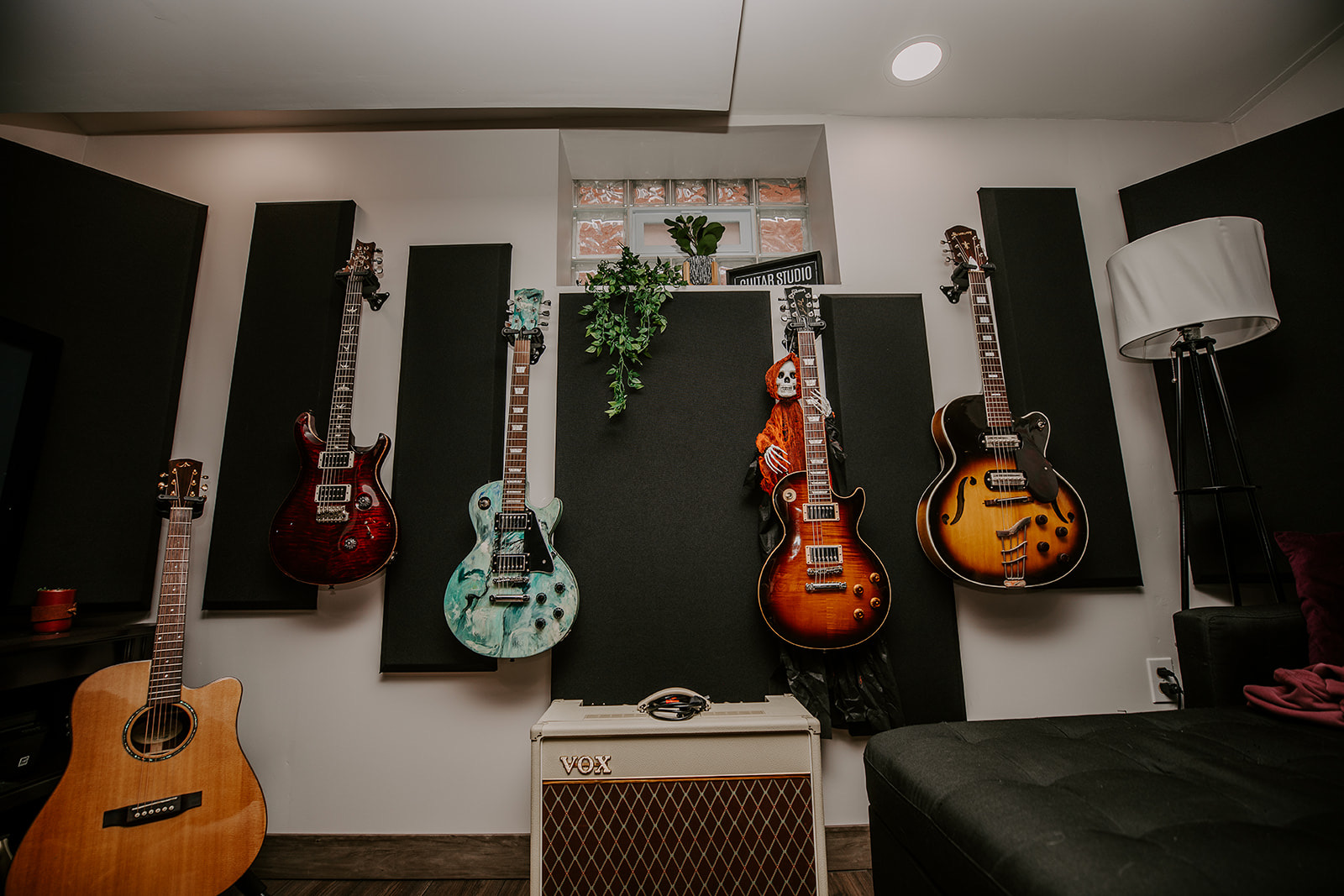 Five guitars hanging on a wall with black acoustic panels, a small window, and a white speaker in the center, with a black couch nearby.