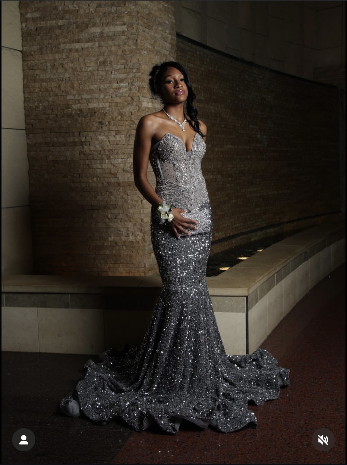 Woman in a sparkly, strapless, floor-length gown with a train, standing against a curved wall, holding a small clutch.