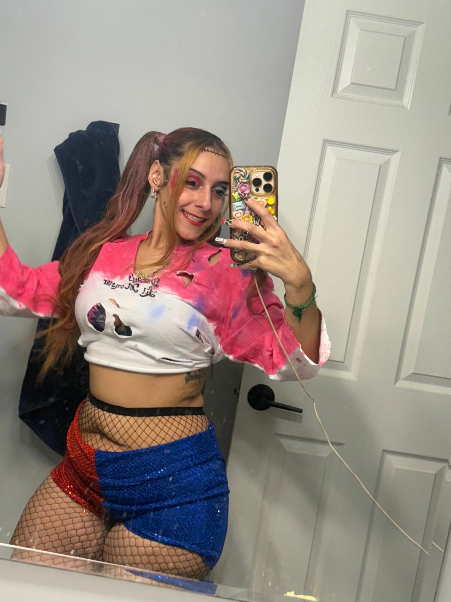 Person taking a mirror selfie, wearing a pink jacket, white shirt, blue shorts, and fishnet stockings, with a phone in hand.