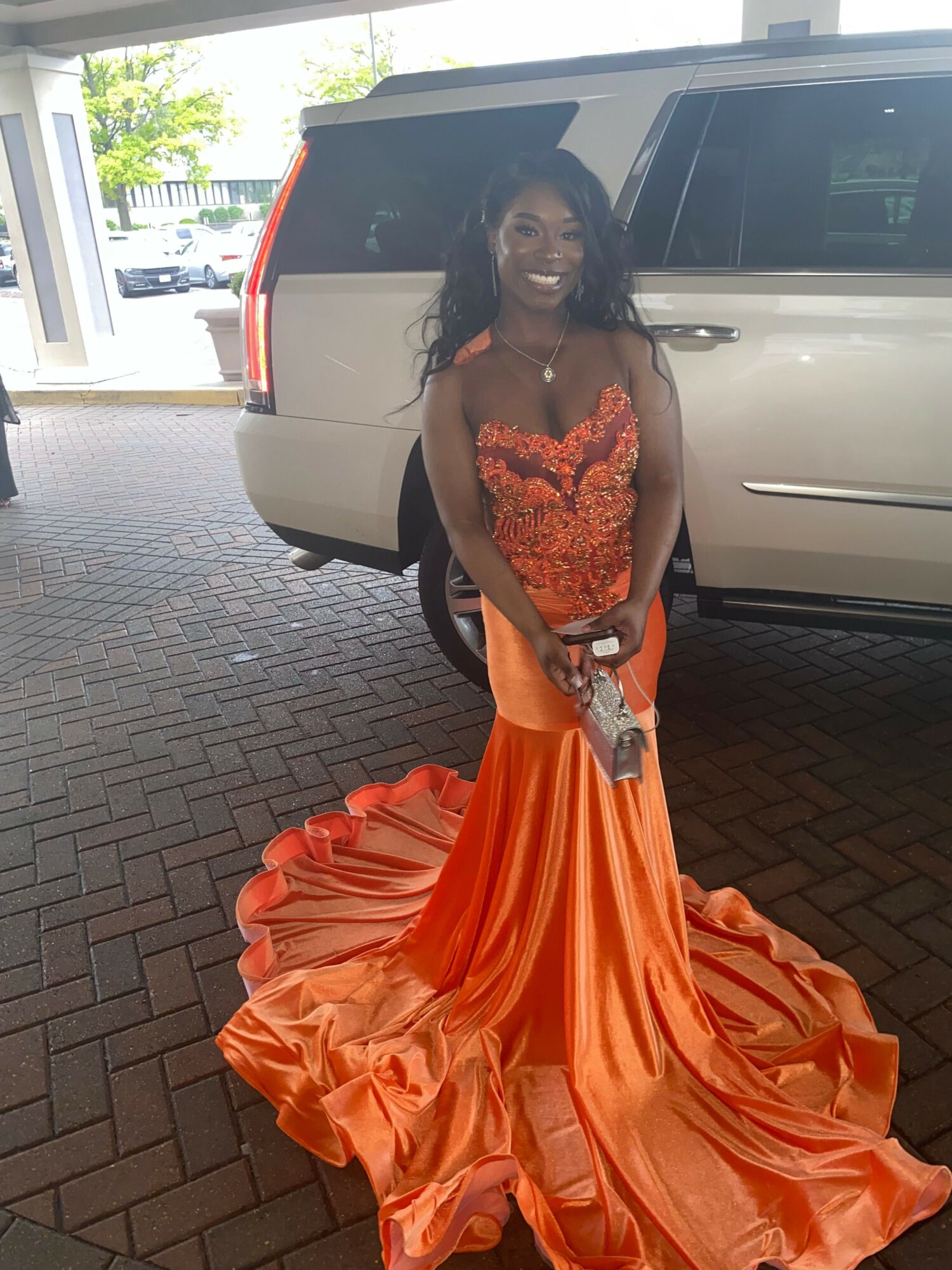 Woman in an orange gown standing outdoors near a vehicle, with trees and buildings in the background.