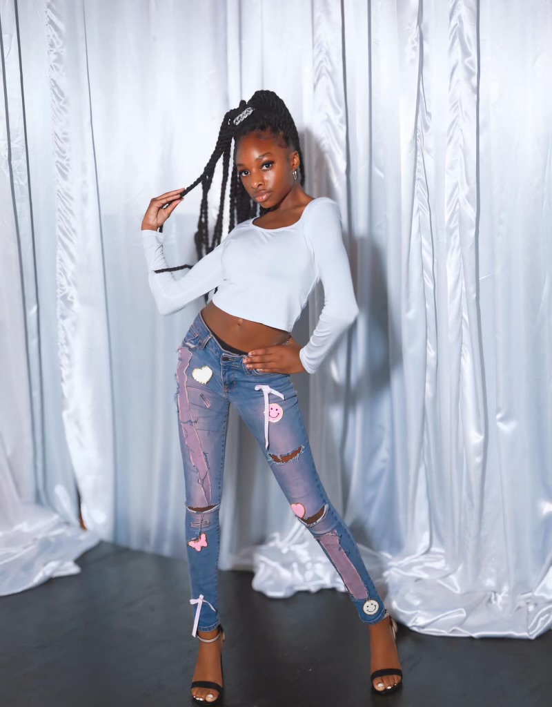 Young woman with long braided hair poses in front of white curtains, wearing a white crop top and ripped jeans with patches.