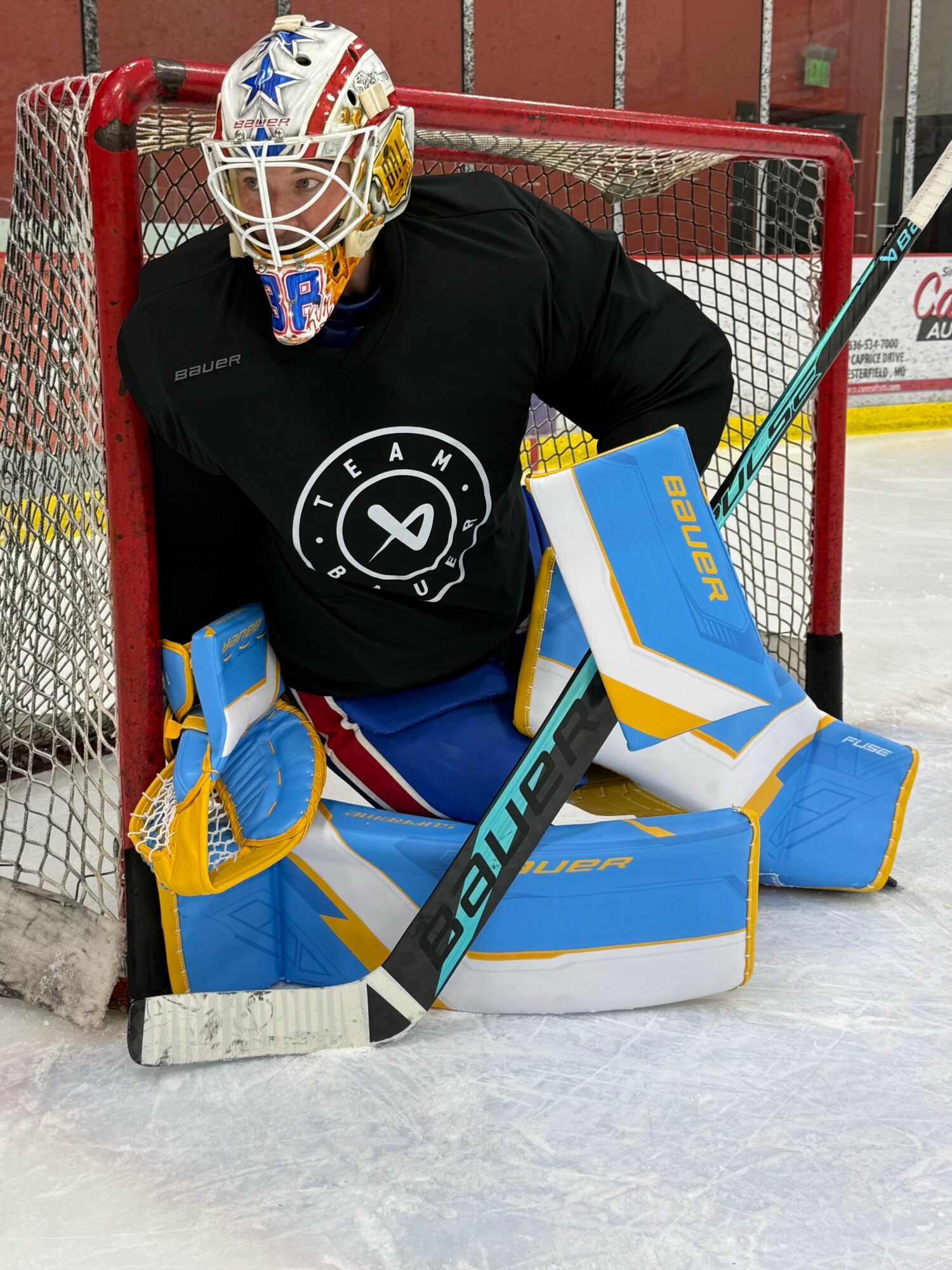 Hockey goalie in black jersey, crouched in front of goal, wearing blue and yellow pads and helmet, holding a hockey stick.