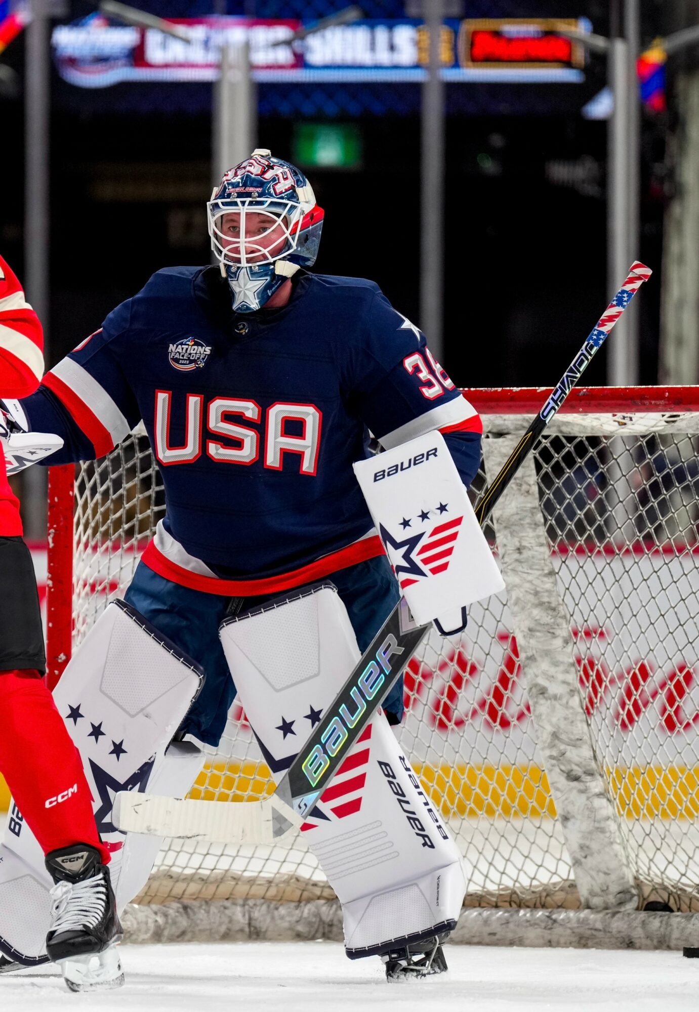 Ice hockey goalie in USA jersey stands in front of goal, holding a stick, with a player nearby on ice.