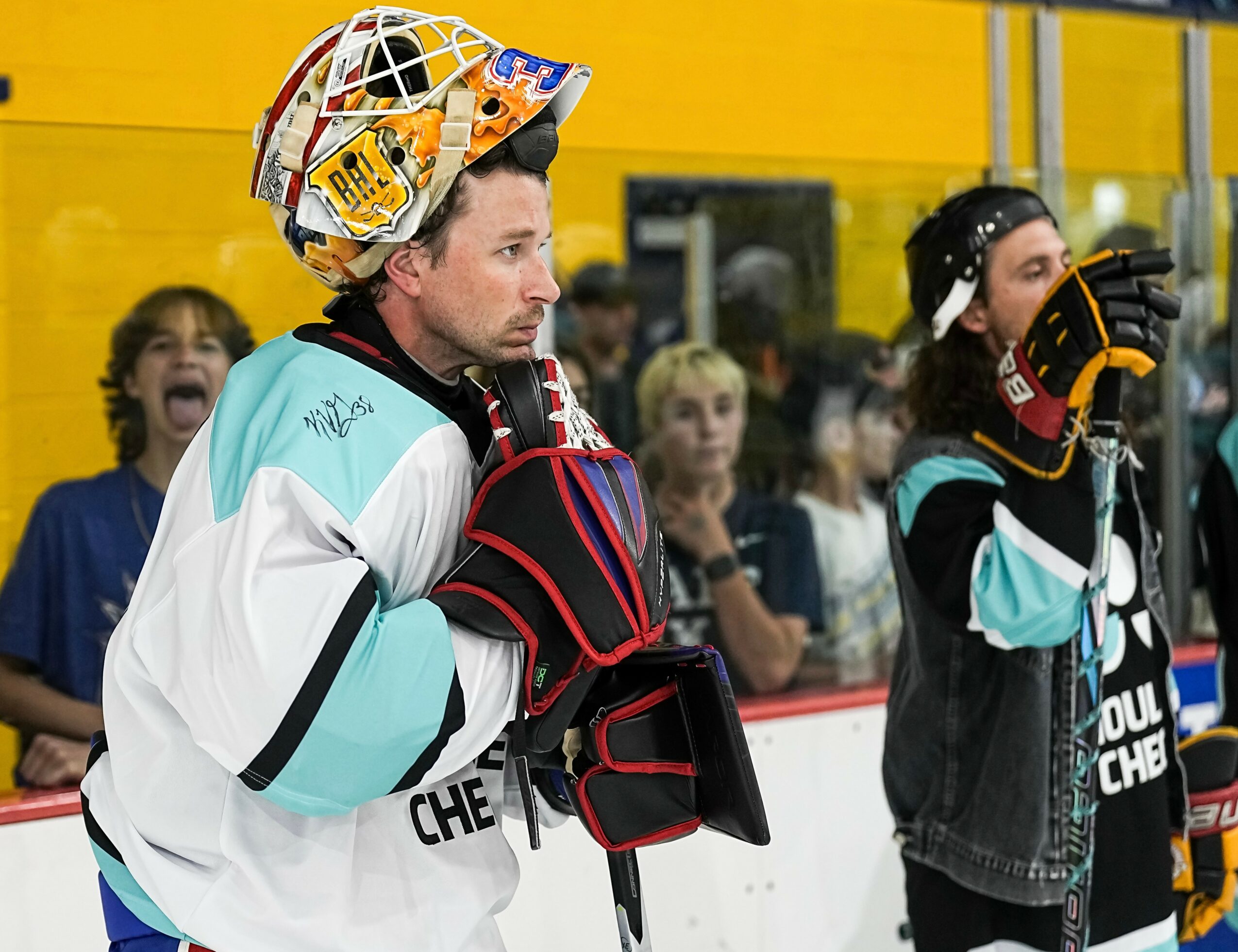 Hockey players in gear on the ice, with spectators in the background, one player adjusting gloves, others watching.