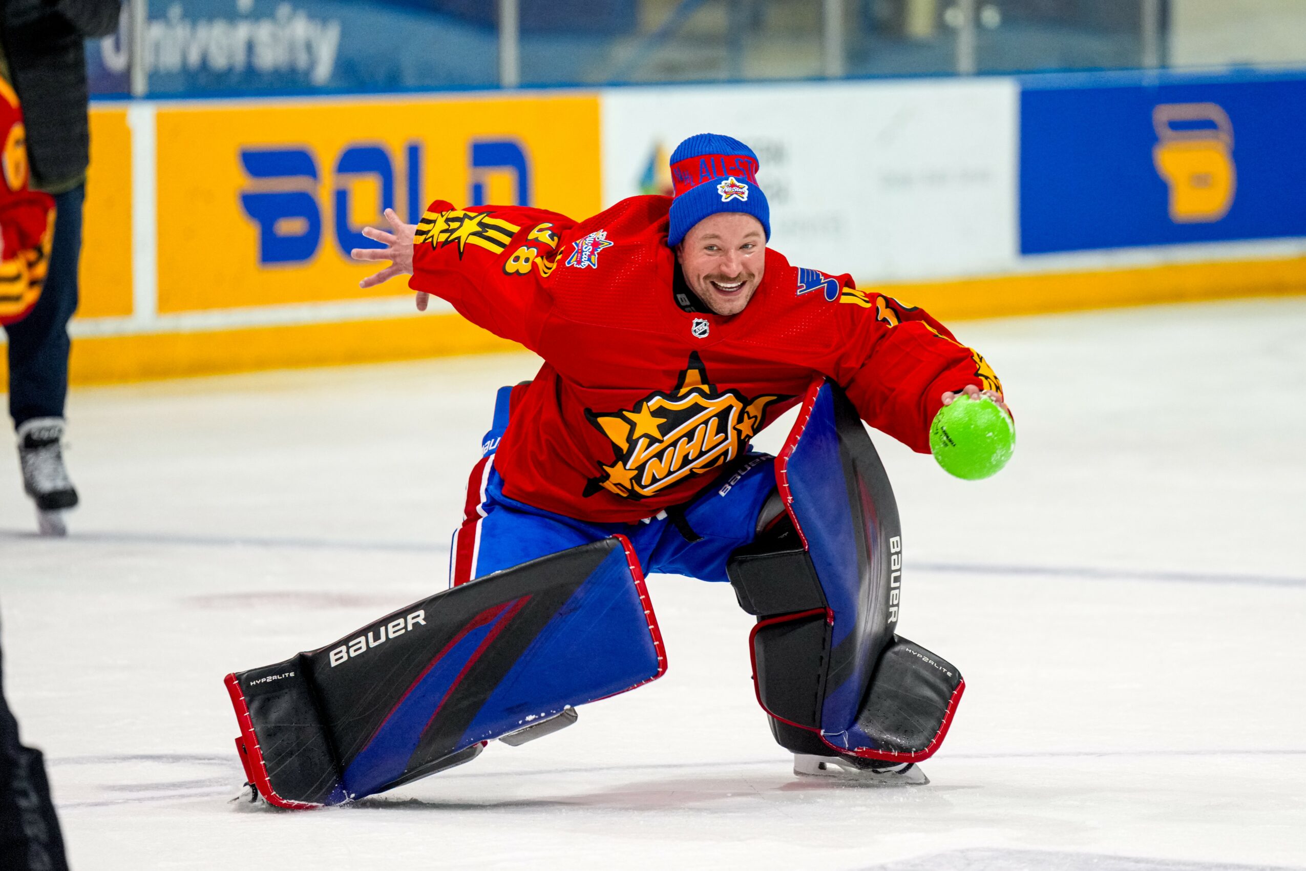A hockey goalie in red uniform crouches on ice, smiling, with a green puck in hand, wearing protective gear.