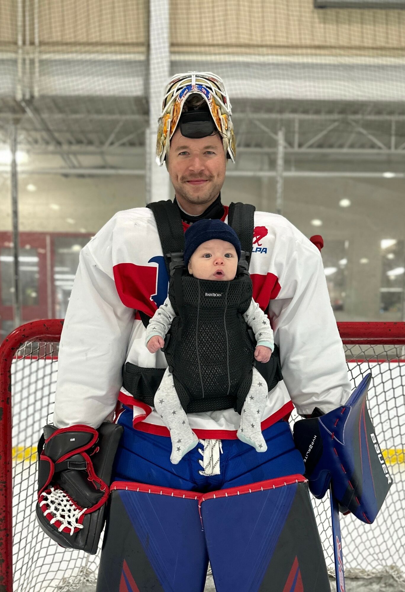 Hockey player in uniform holding a baby in a carrier, standing on ice rink, with red and blue equipment, smiling.